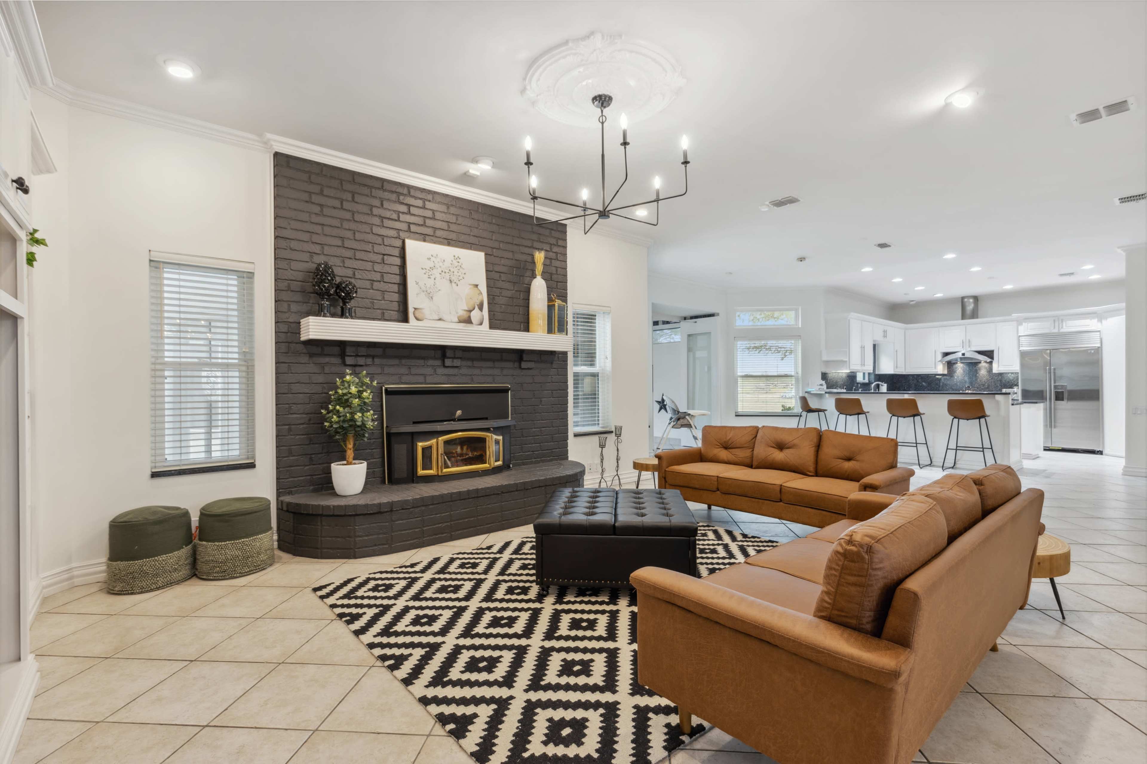 The image shows a modern living room featuring a brown leather sofa, a black upholstered coffee table, and a fireplace surrounded by dark brick, with a patterned rug on the tiled floor.