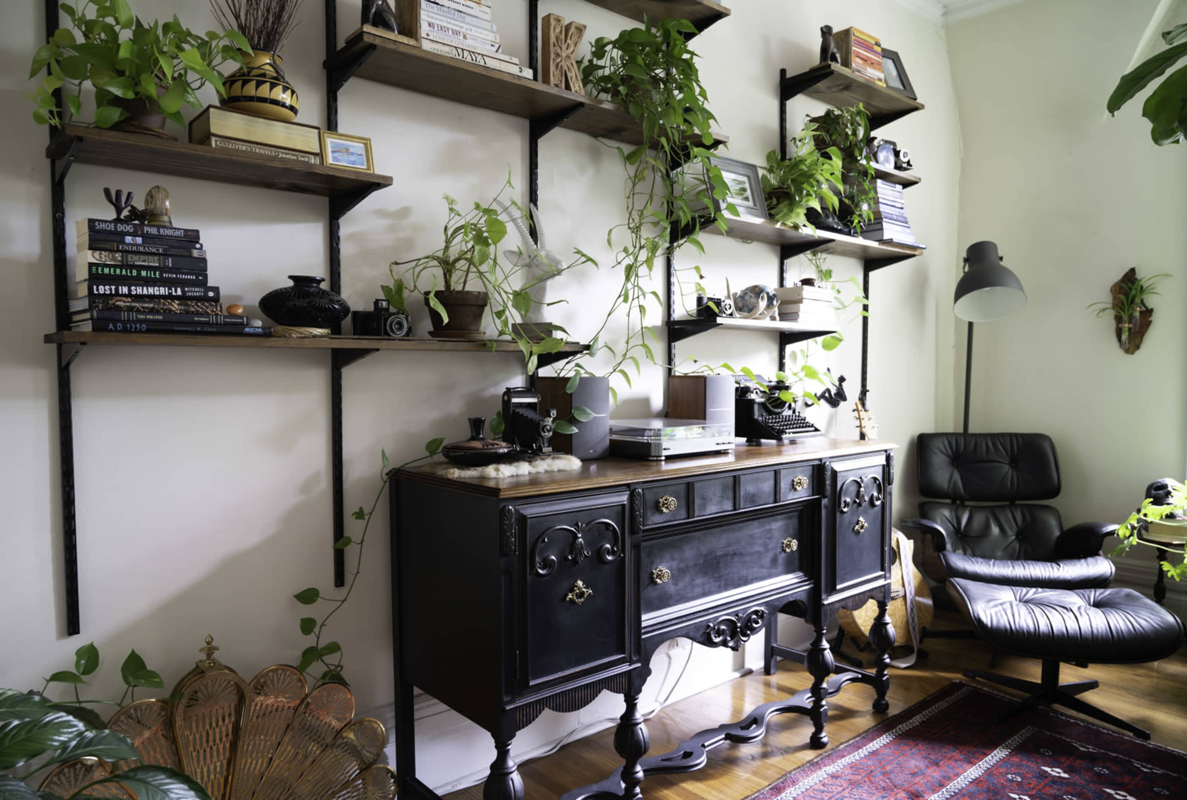 A corner of a room featuring a black sideboard, shelves adorned with books and plants, and a modern black armchair.
