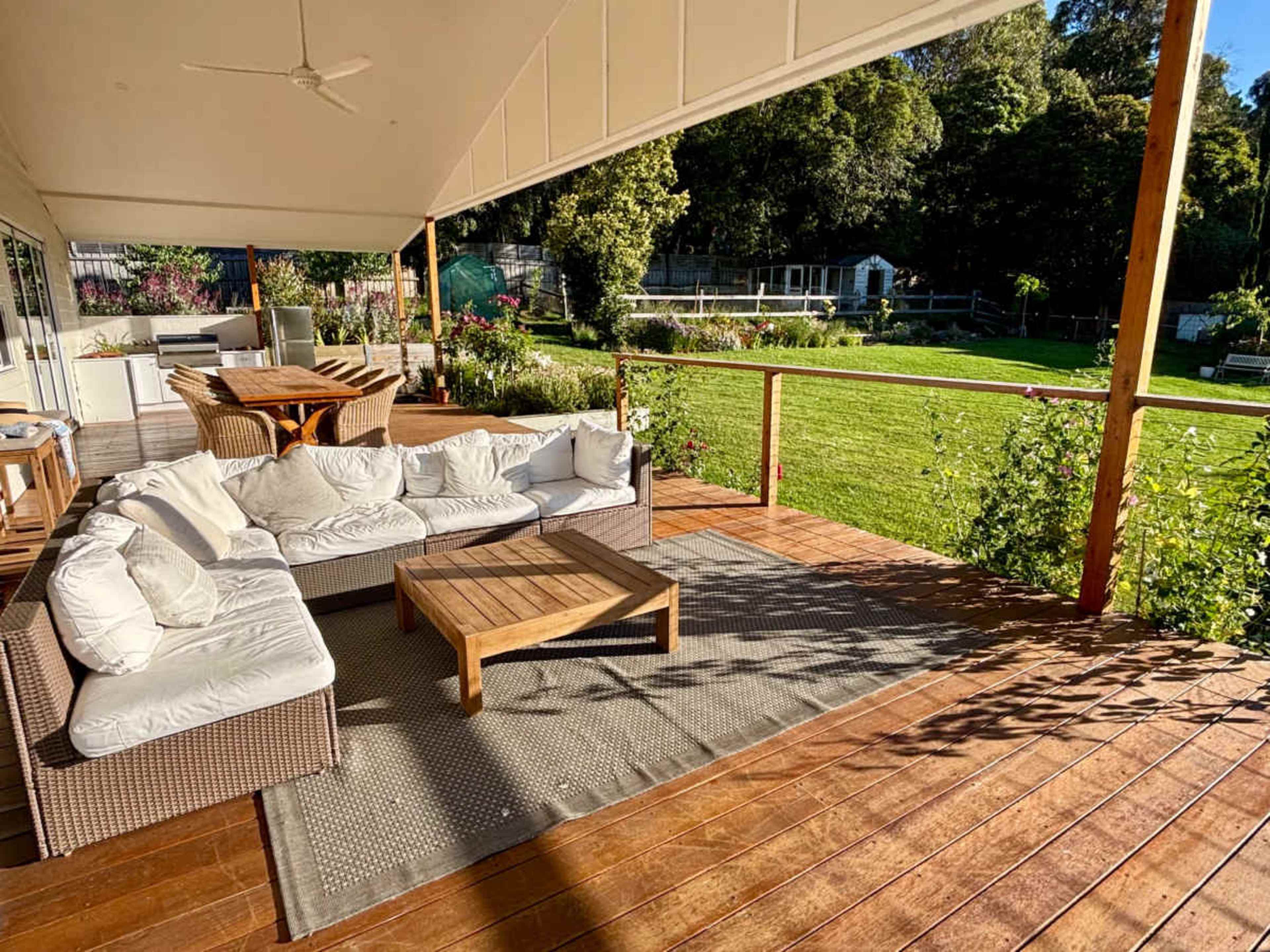 An outdoor seating area features a large sectional sofa, wooden coffee table, and dining set overlooking a green lawn and garden.