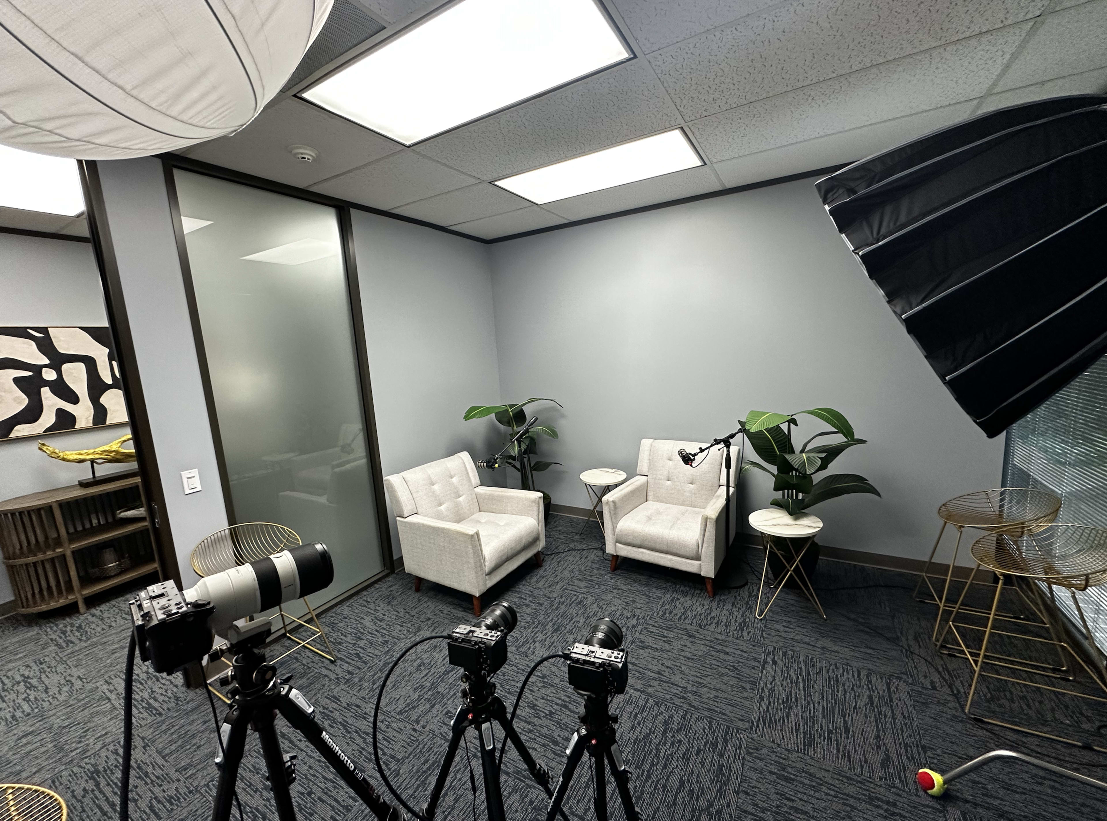 A minimalist recording studio featuring two white chairs, a microphone setup, and several cameras positioned on tripods.
