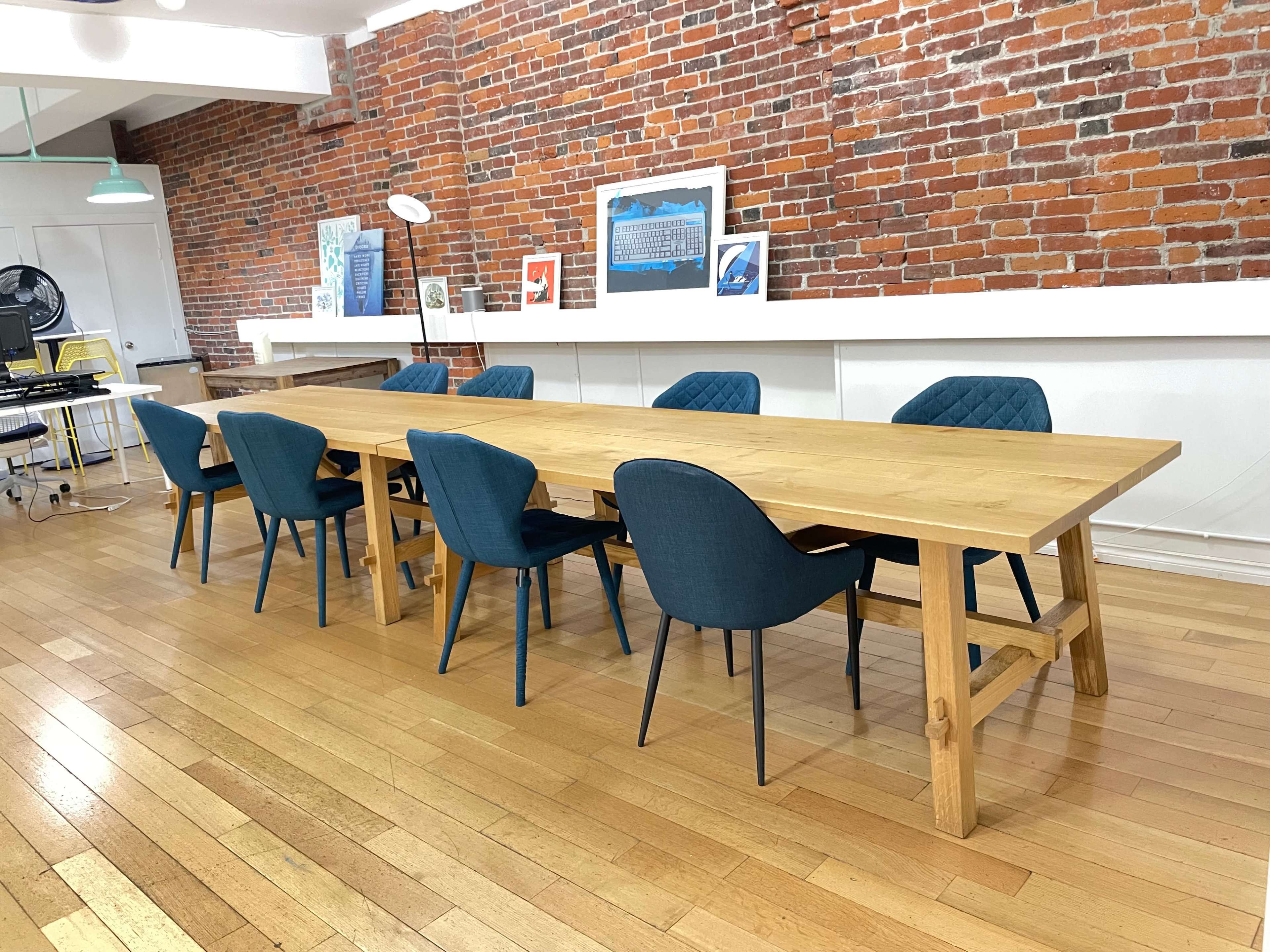 A long wooden table surrounded by six upholstered chairs is positioned in a room with exposed brick walls and wooden flooring.