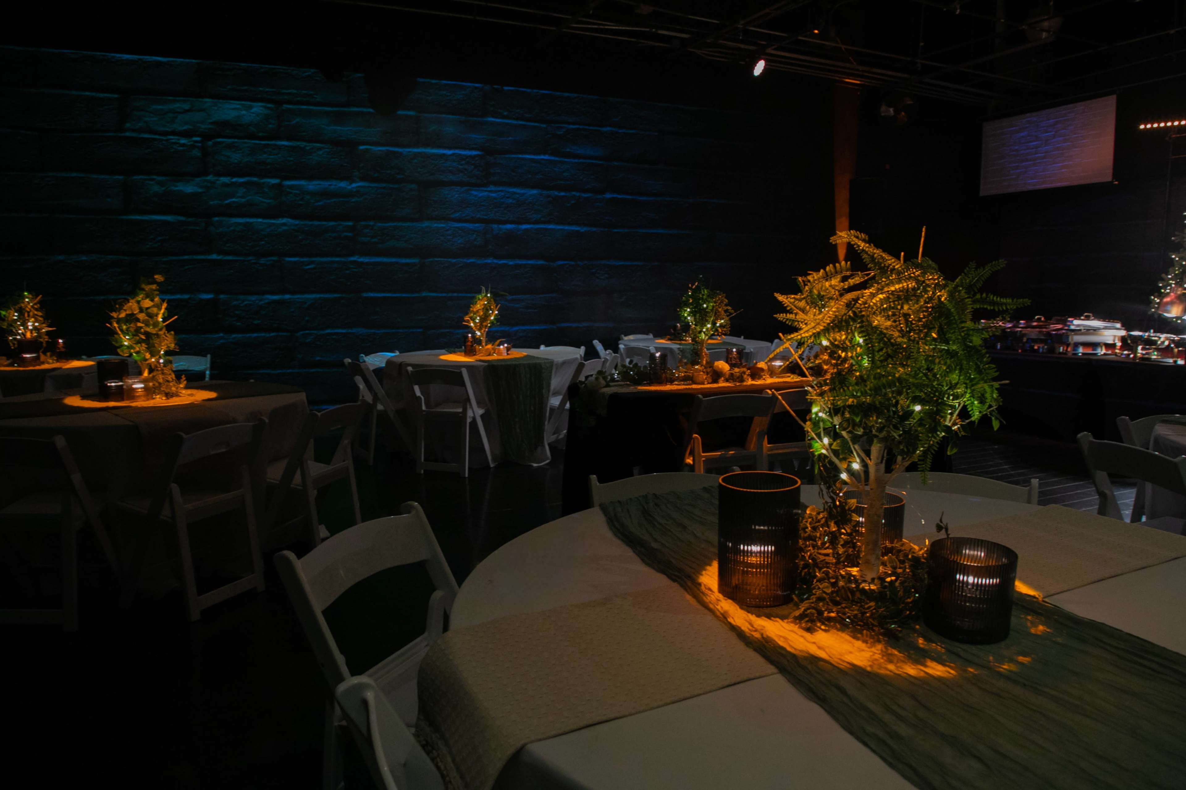 The image shows a dimly lit event space with round tables, each adorned with greenery and warm lighting against a textured blue wall.