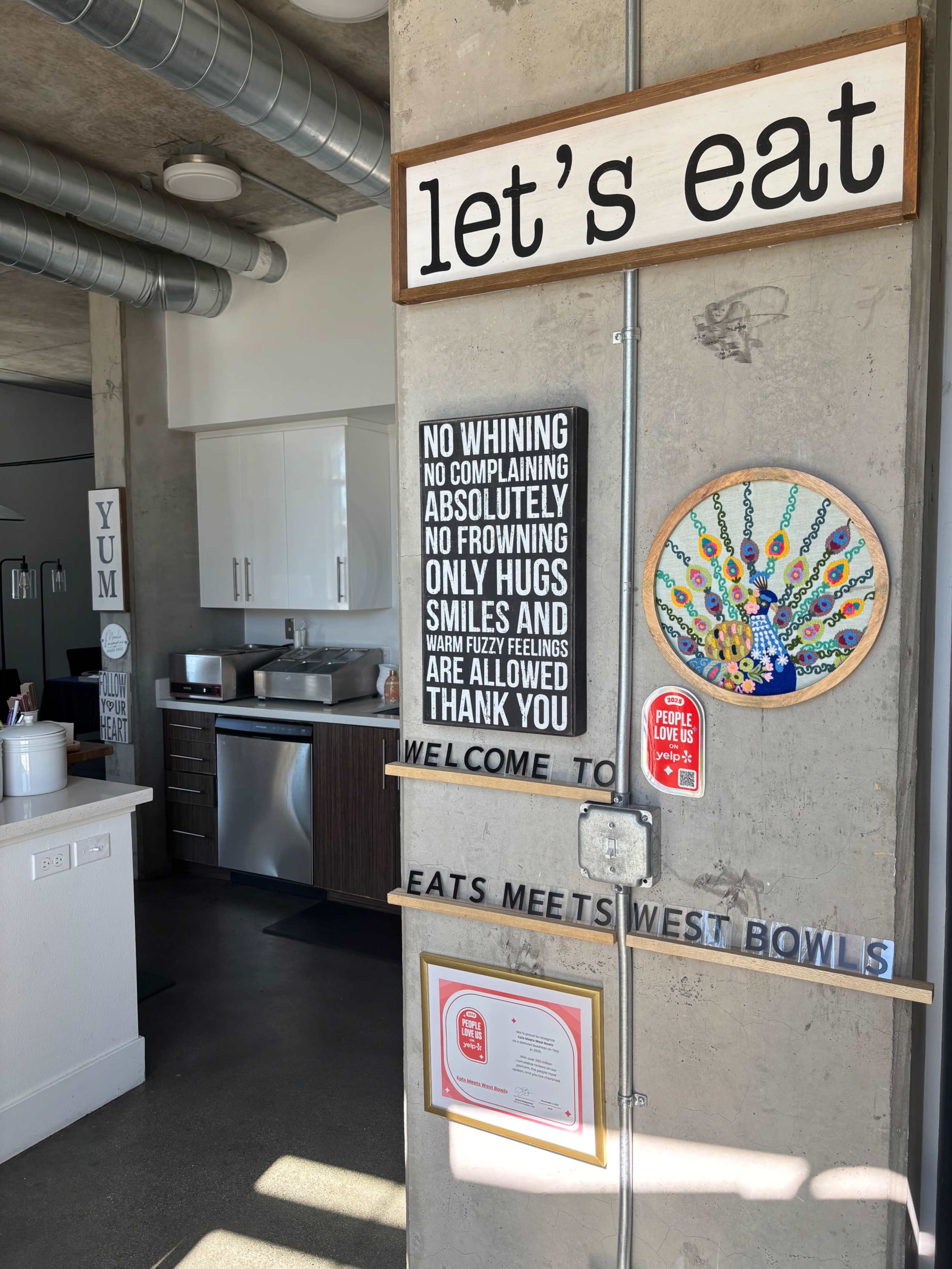 The image shows a dining area with a concrete wall featuring signs that read "let's eat," "NO WHINING," and "WELCOME TO EATS MEETS WEST BOWLS," alongside a colorful peacock art piece.