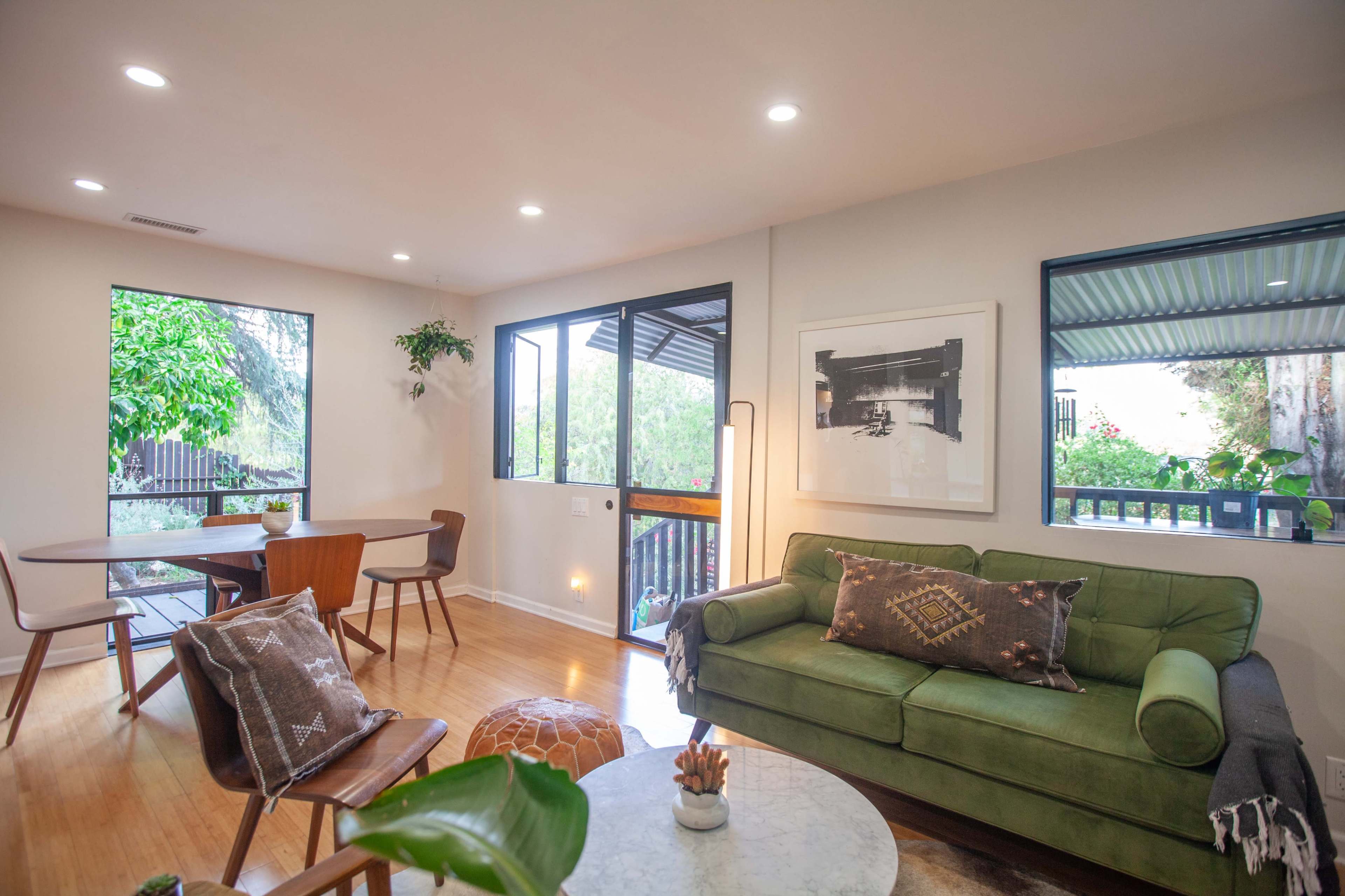 A bright living room features a green sofa, round coffee table, and a dining area with a wooden table near large windows overlooking a garden.
