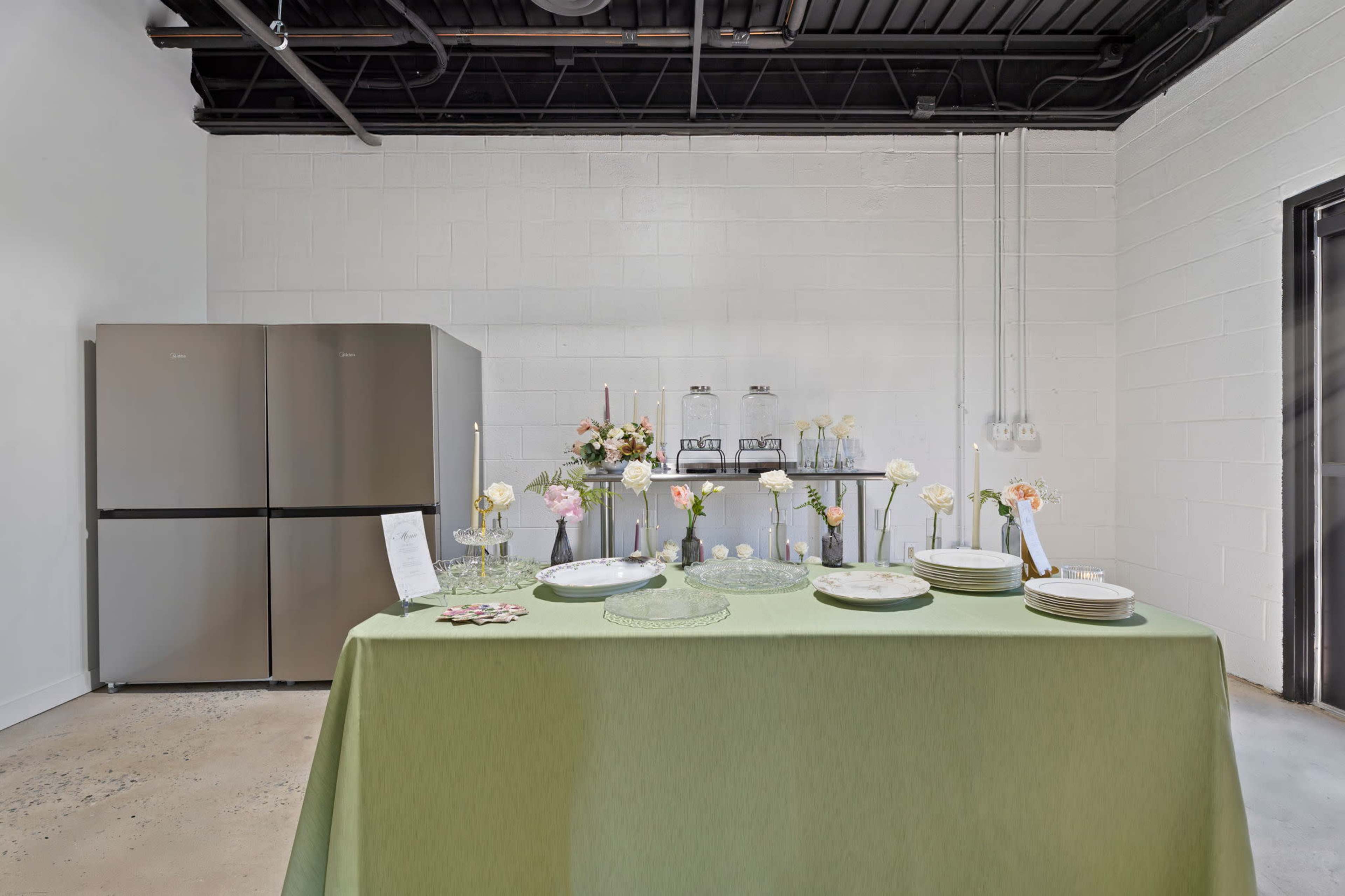 The image shows a banquet setup featuring a green tablecloth, plates, glassware, a floral arrangement, and a silver beverage station in a room with white walls and a gray refrigerator.