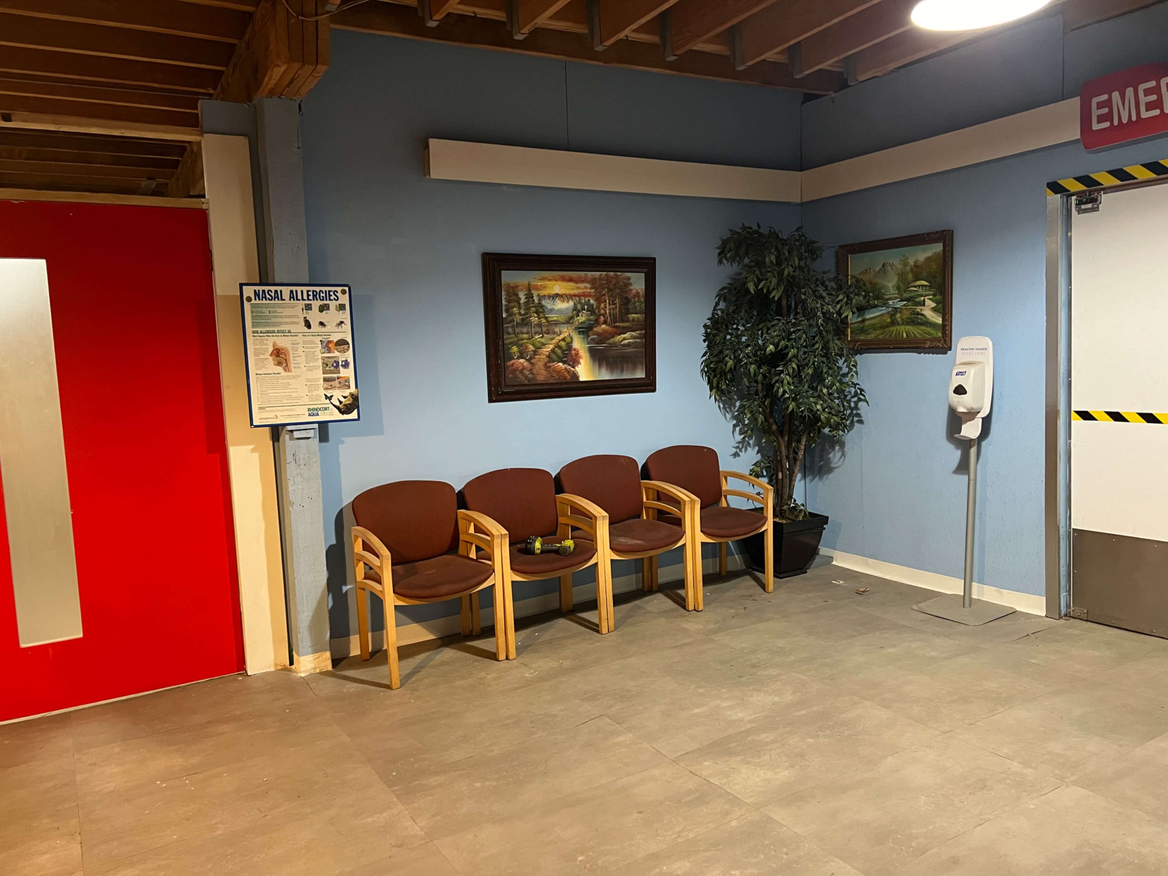 A waiting area with three wooden chairs, a potted plant, two framed pictures on the wall, and a wall-mounted hand sanitizer dispenser in a blue and white room with a red emergency exit door.