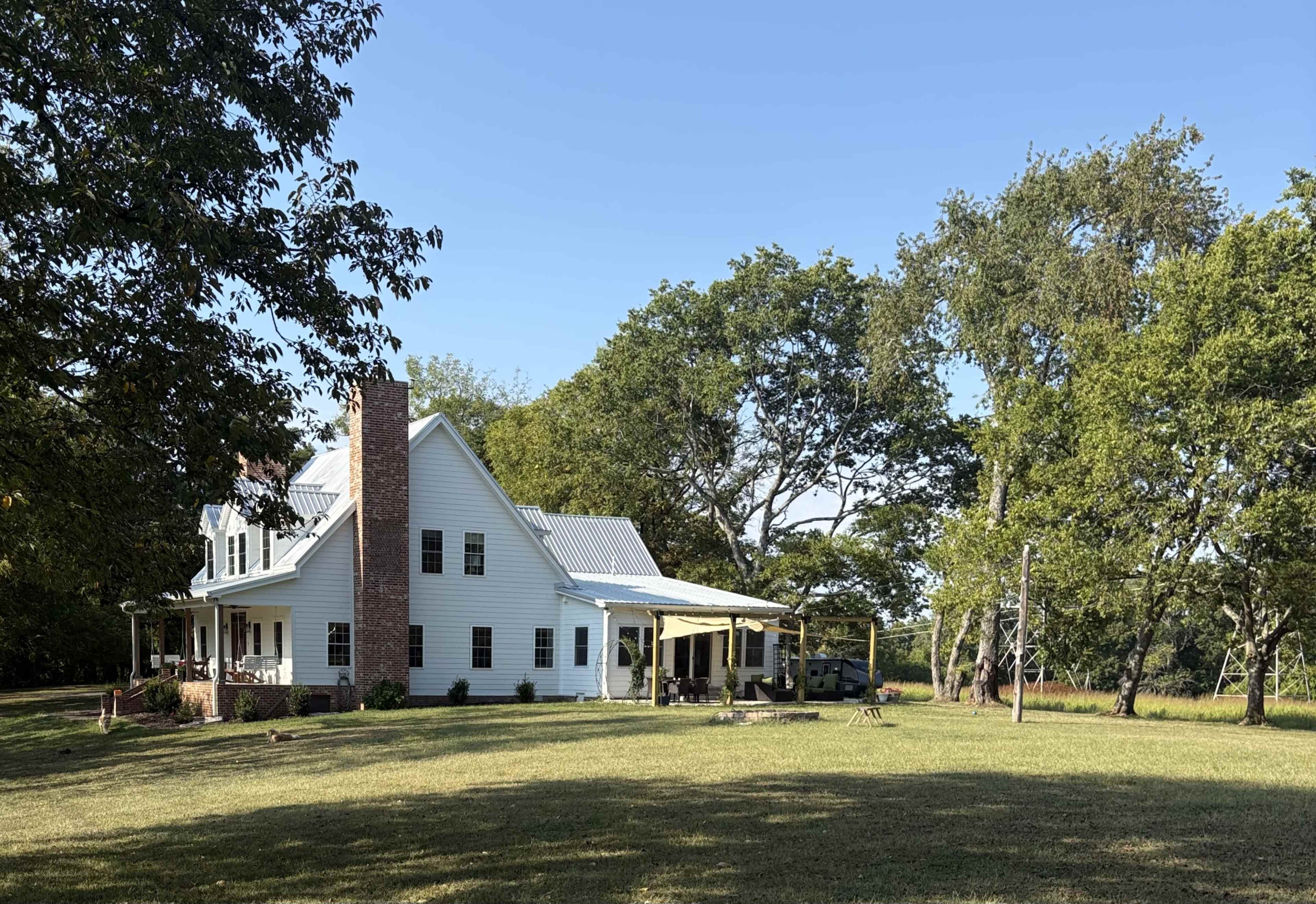 A two-story white farmhouse with a porch and a chimney stands in a grassy area surrounded by trees under a clear blue sky.