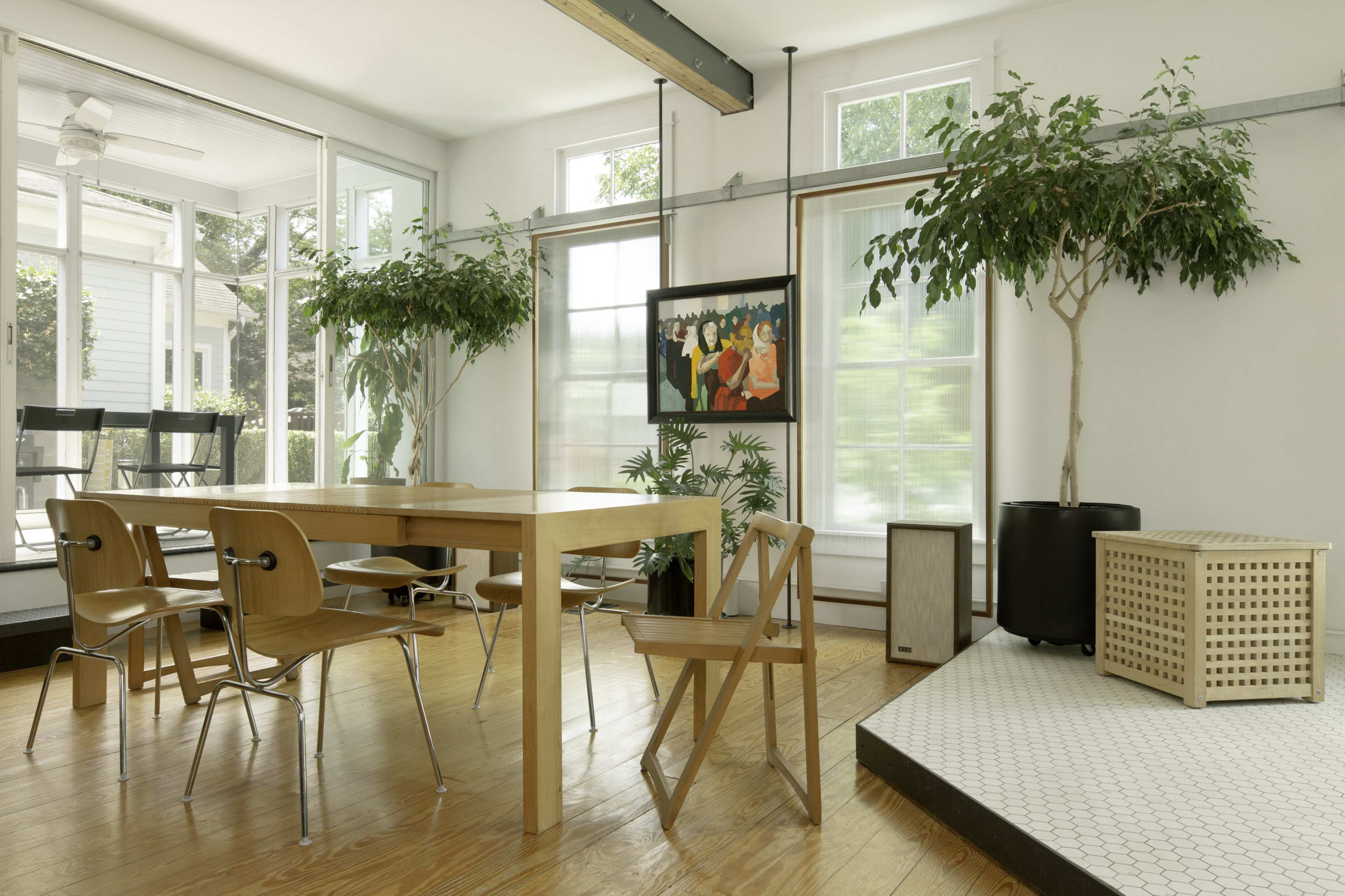 A well-lit dining area featuring a wooden table surrounded by chairs, with large windows, potted plants, and a painting on the wall.