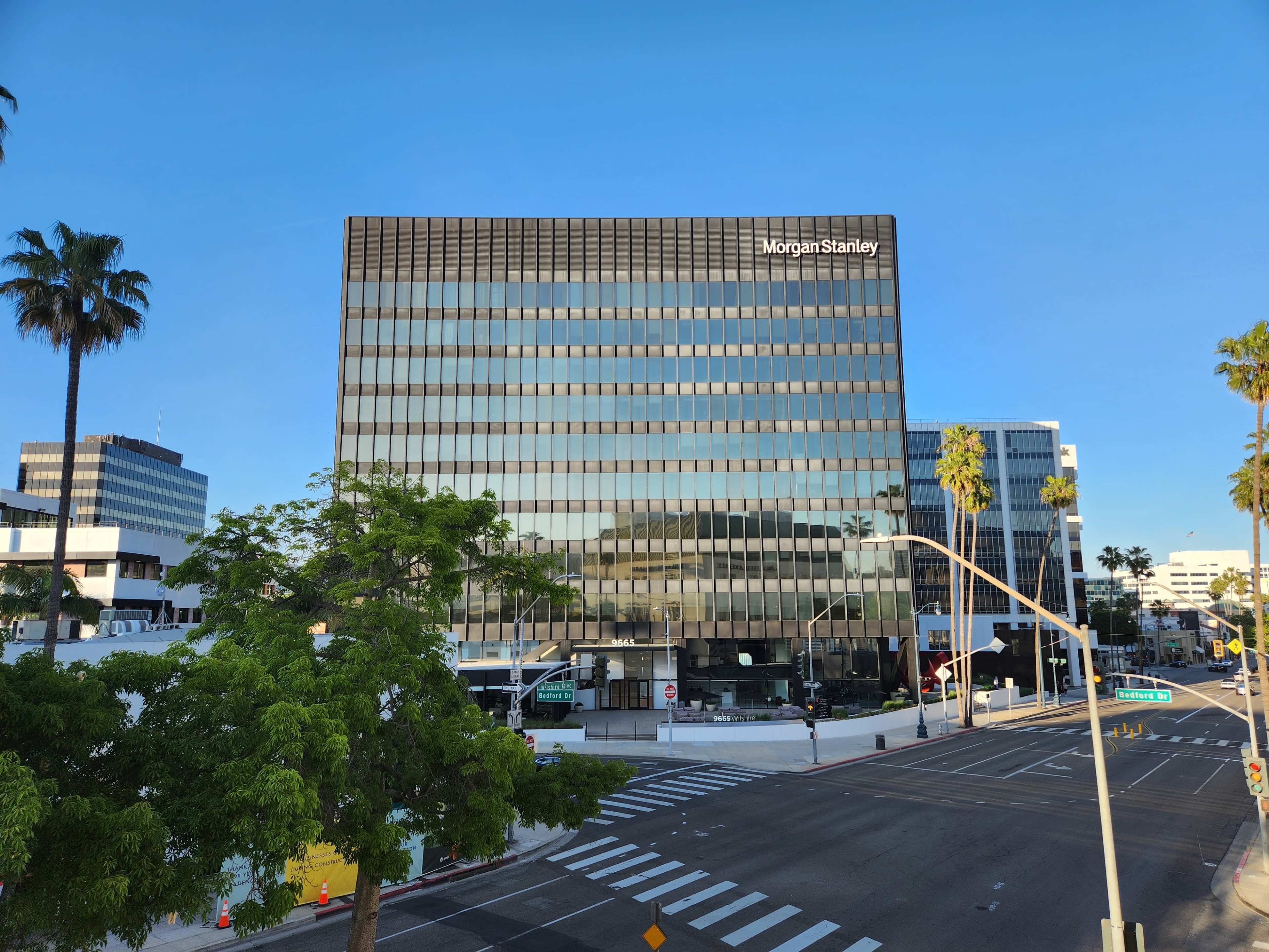 A modern office building with large glass windows, featuring the Morgan Stanley logo, is located at the intersection of a tree-lined street.
