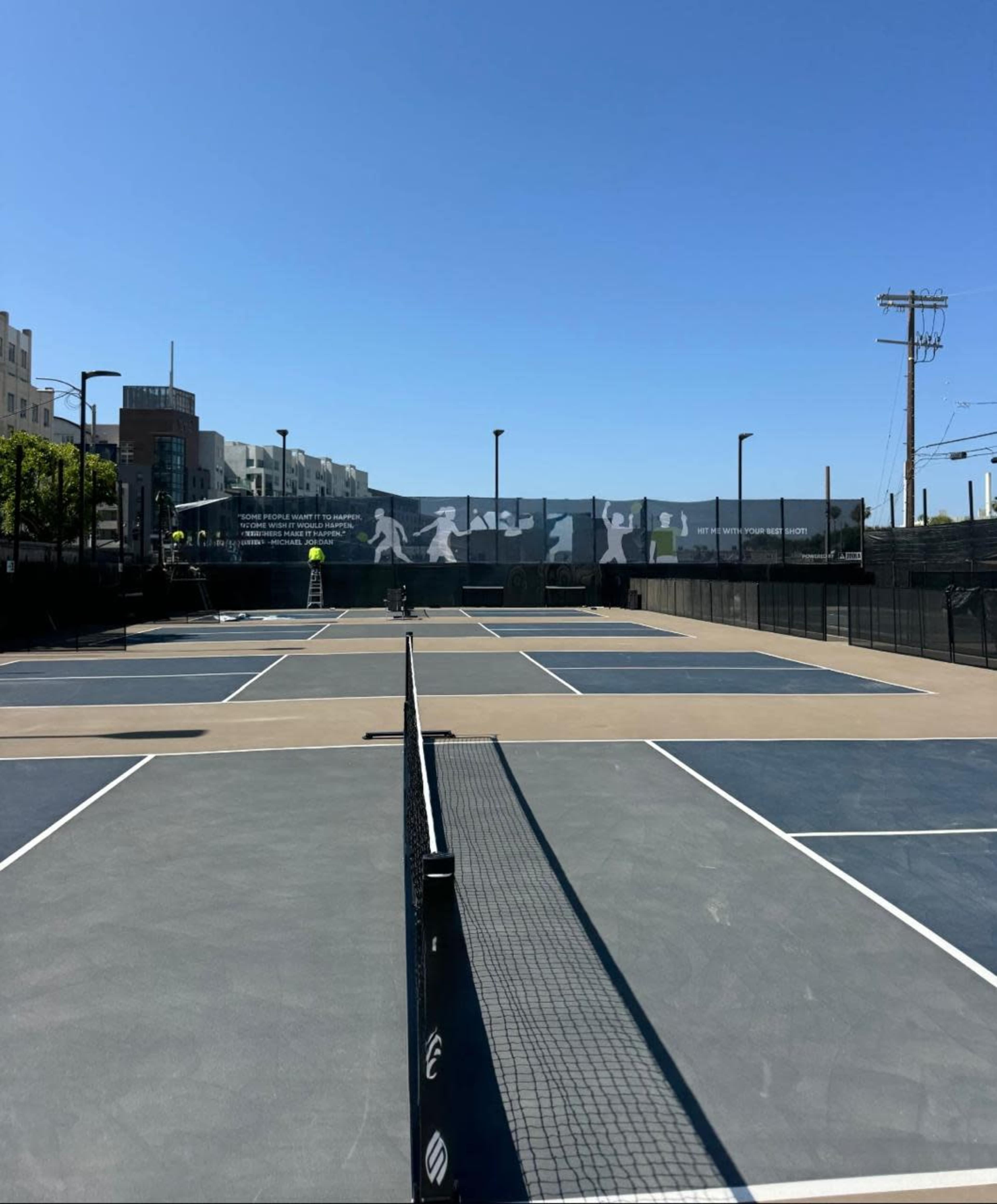 The image shows a series of tennis courts under a clear blue sky with a fence displaying a mural in the background.