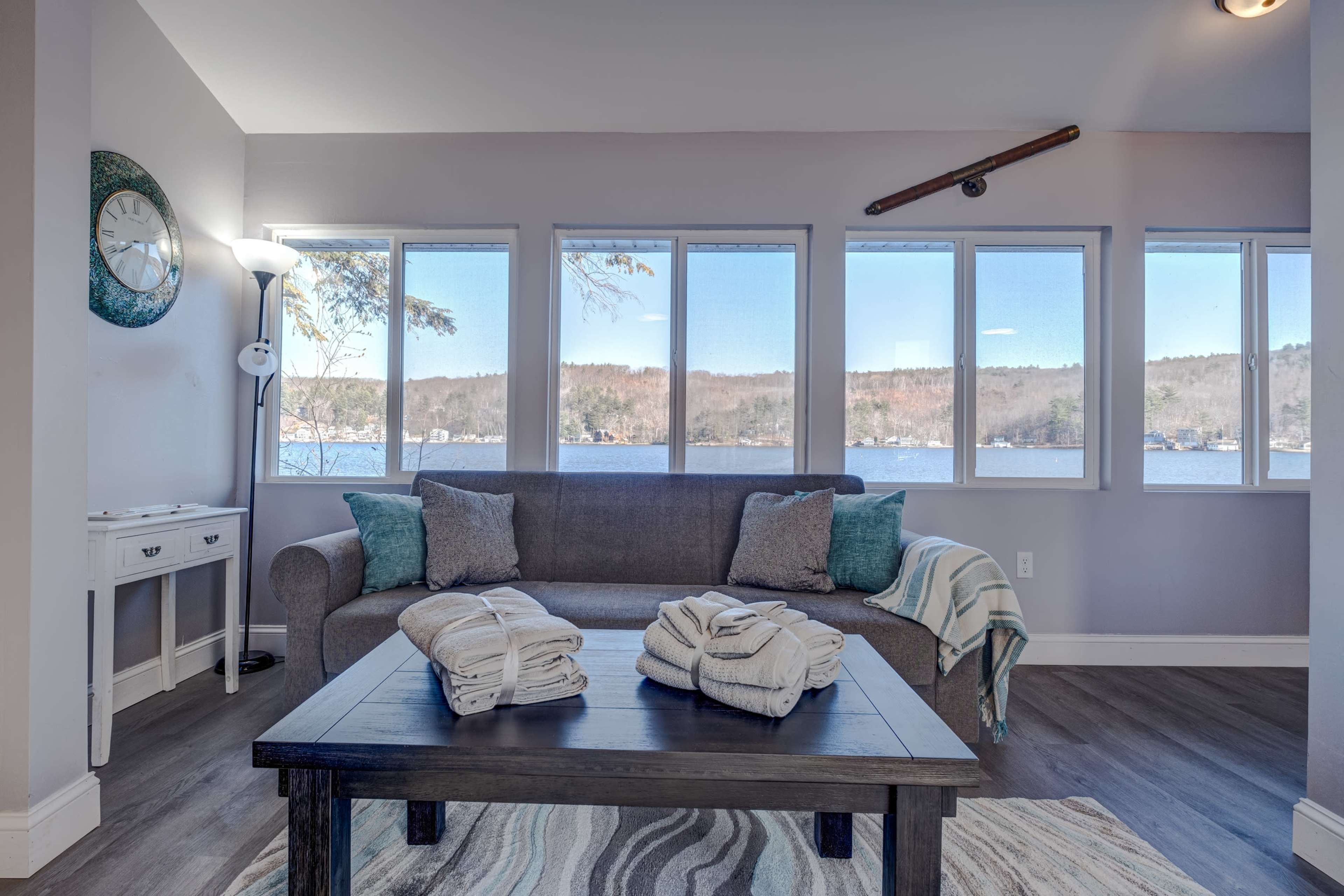 A cozy living room features a gray sofa with folded towels on a black coffee table, overlooking a lake through large windows.