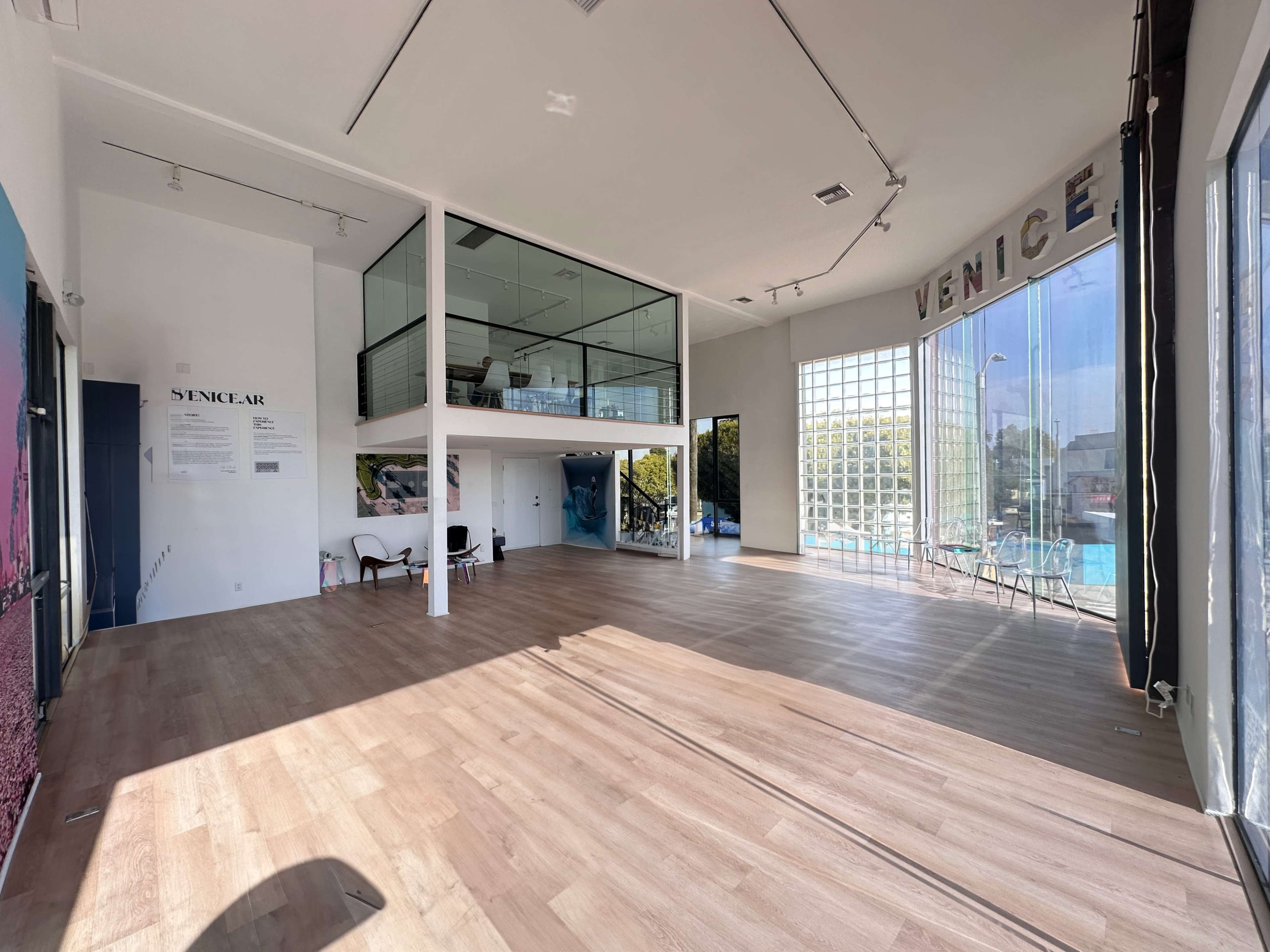 The image shows a spacious art gallery with large windows, a two-level setup, and polished wooden floors.