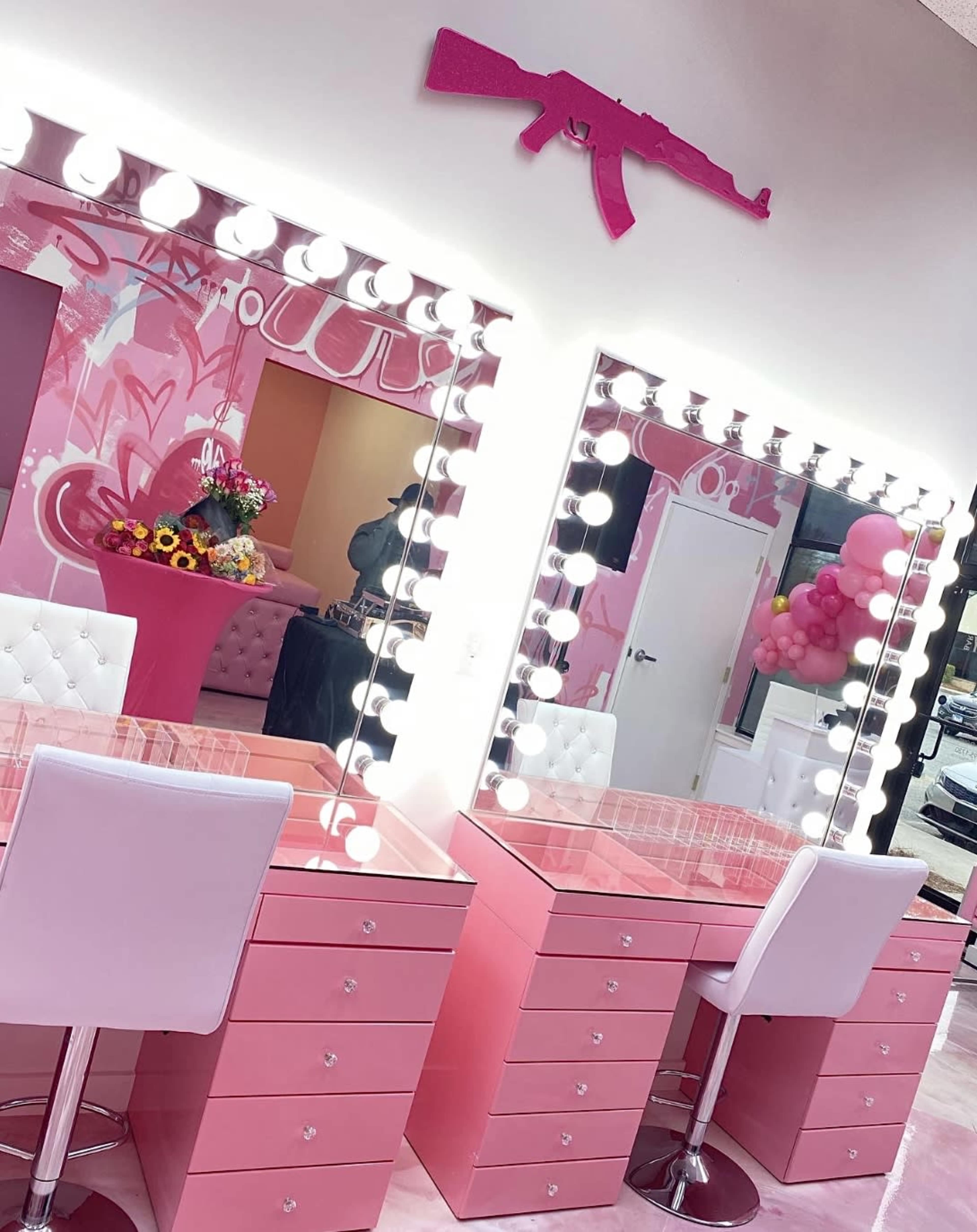 The photo shows a salon area featuring two pink makeup vanities with circular lights and a pink decorative item resembling an assault rifle mounted on the wall above.