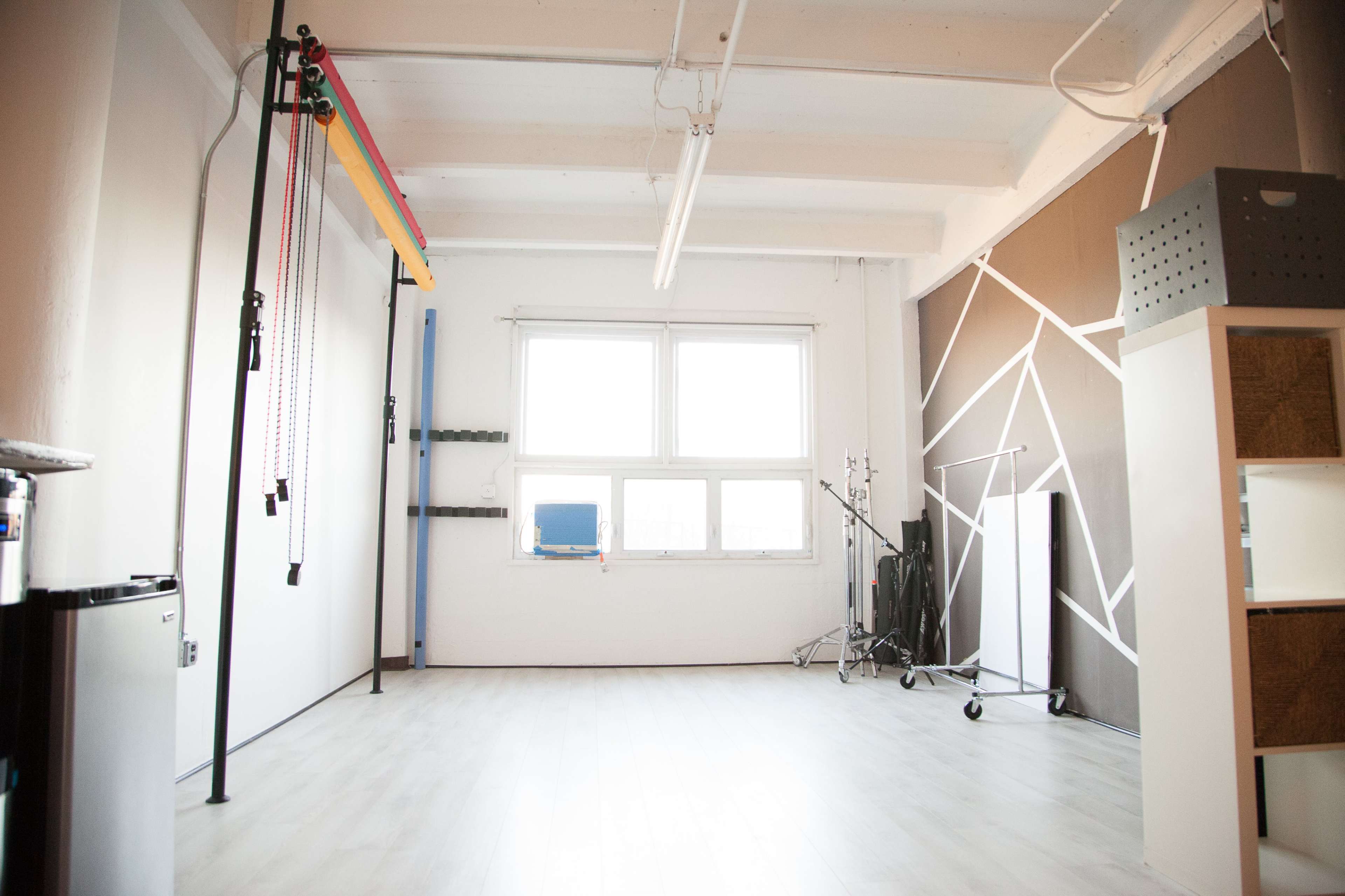 The image shows a spacious, well-lit studio with a white floor, a large window, and various equipment including a backdrop stand and storage units.