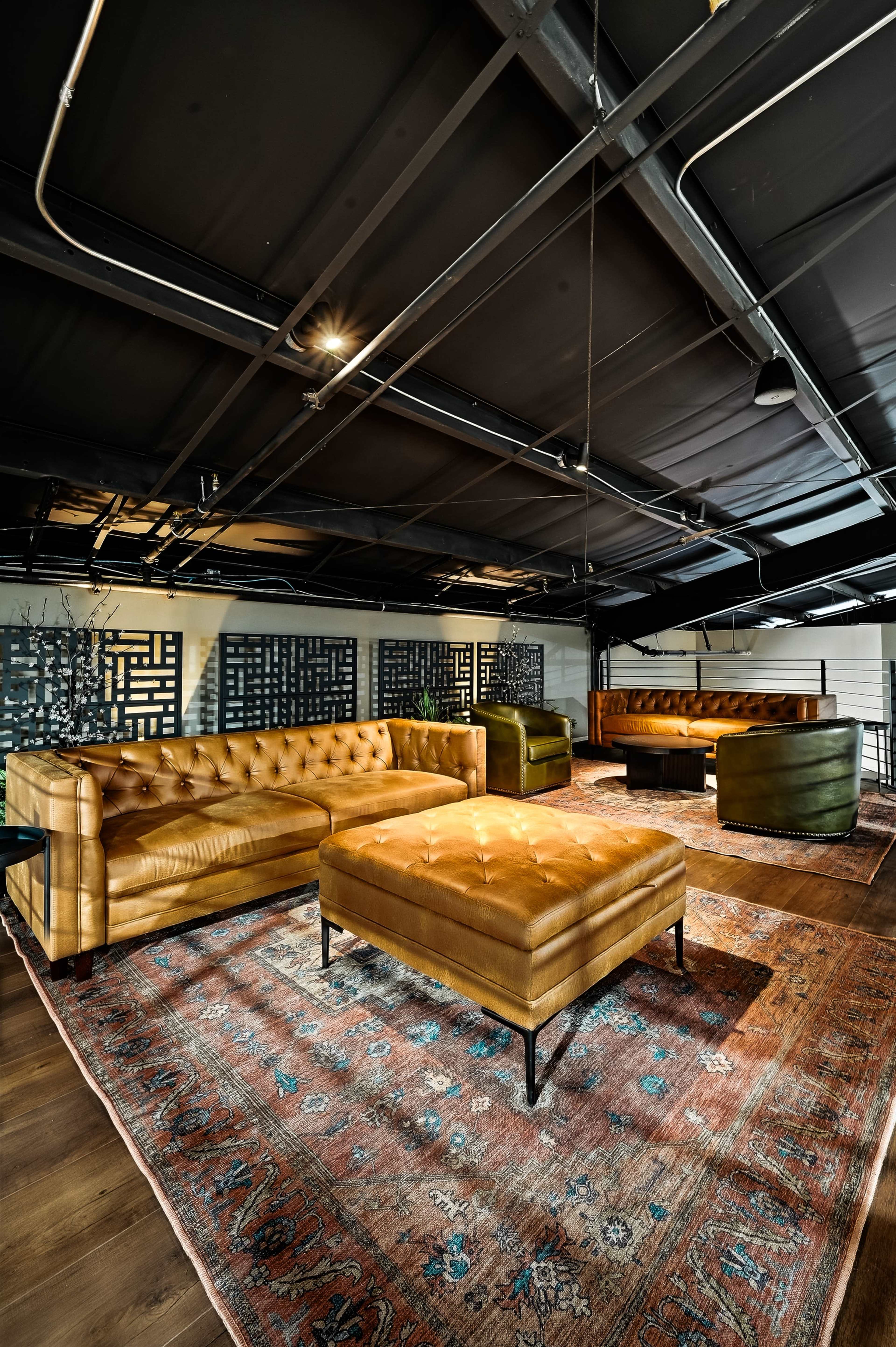 The image shows a modern lounge area with tufted leather sofas, an ottoman, and a patterned rug in a spacious room with dark ceilings.