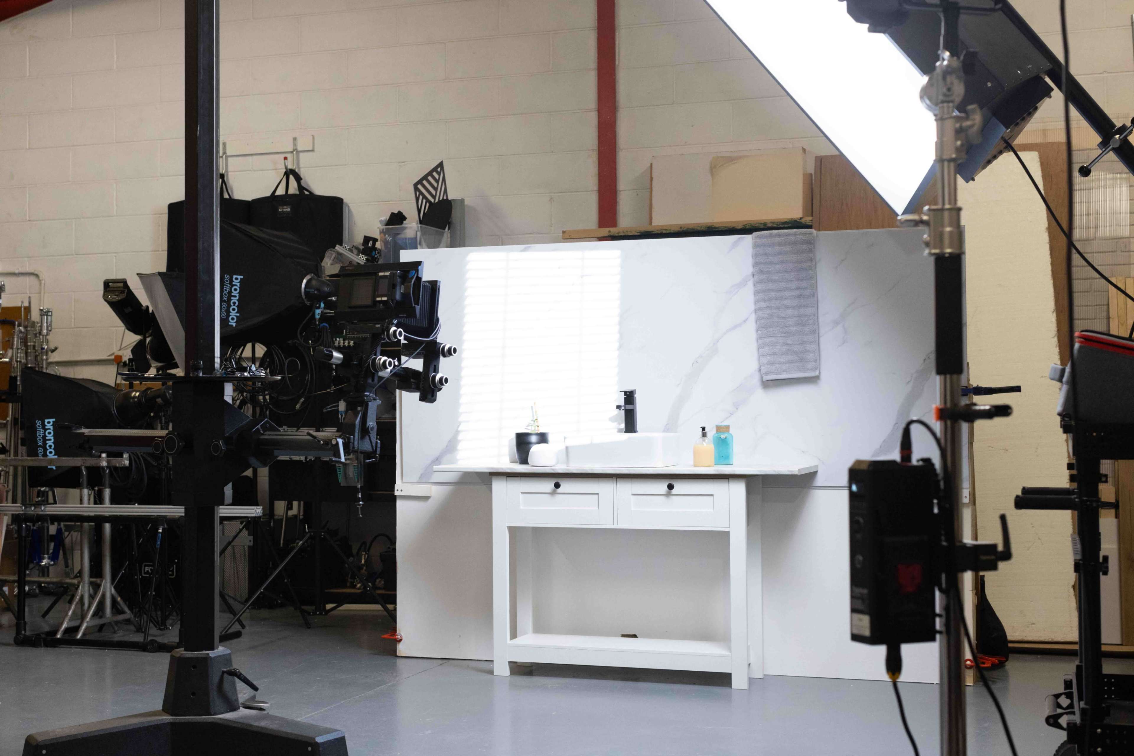 A well-lit studio setup features a white vanity with a sink and two containers, surrounded by camera equipment and studio lights.