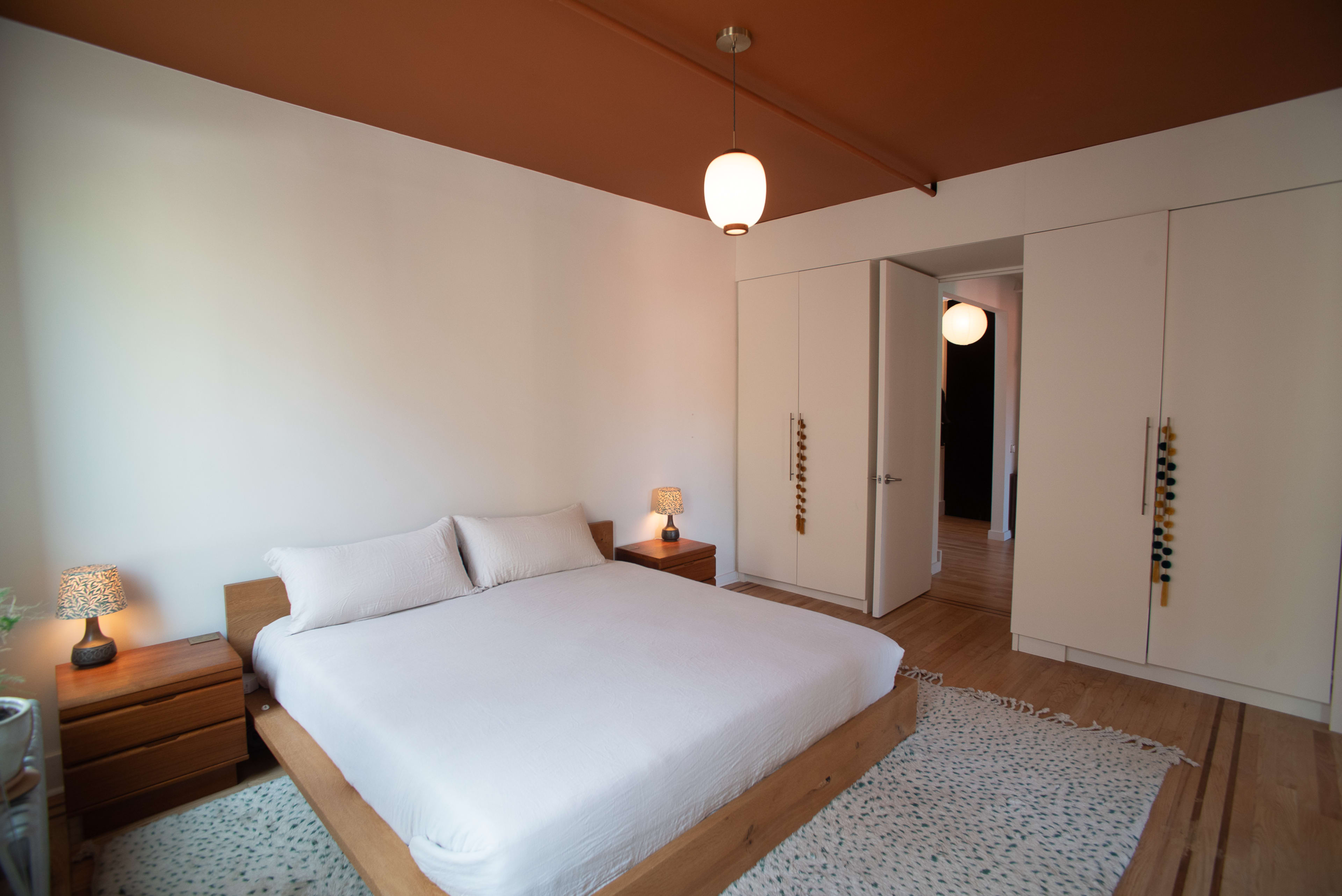 A minimalist bedroom features a wooden bed with white bedding, two bedside lamps, and a light-colored rug, with a closet and doorway visible in the background.