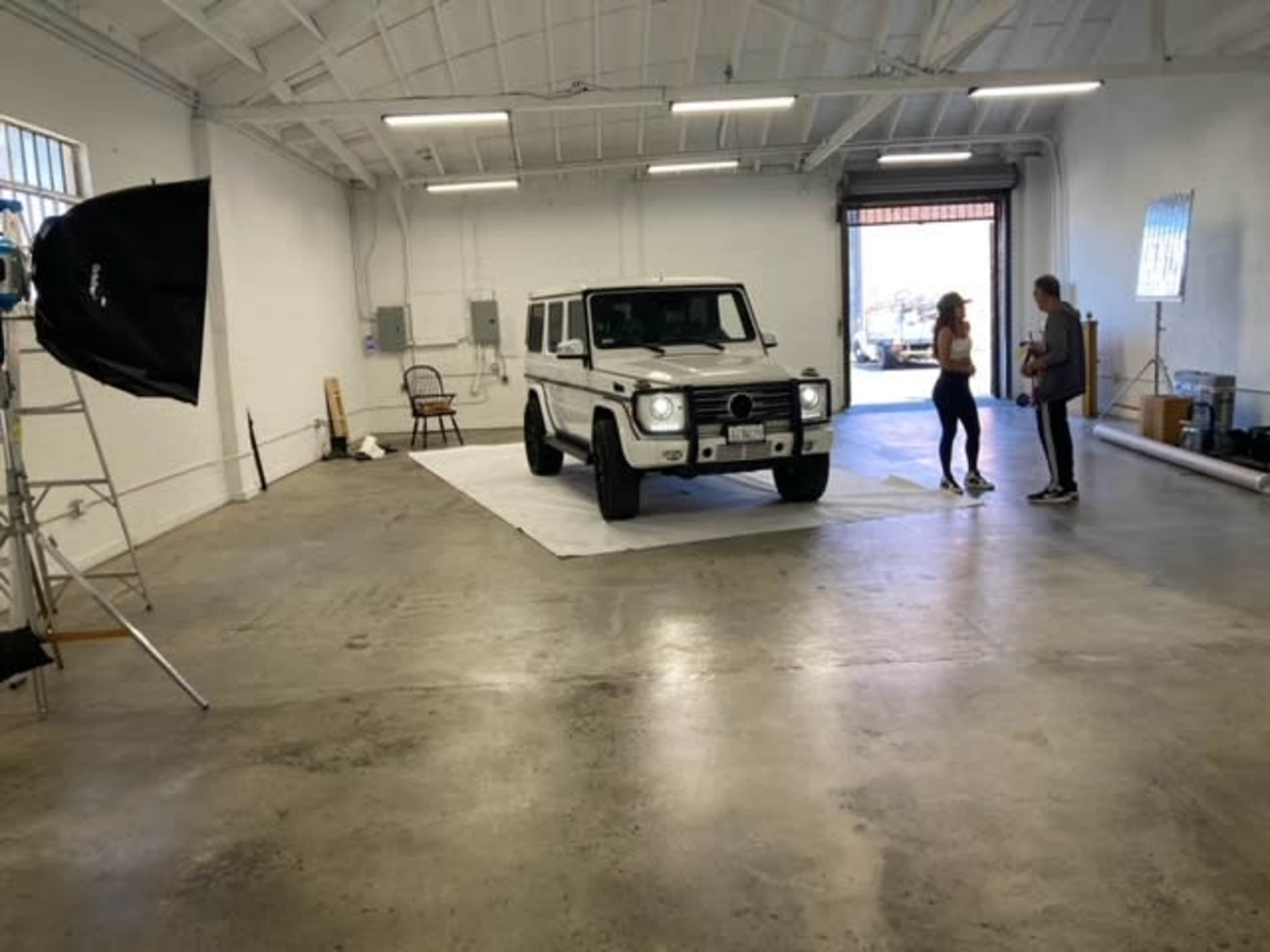 A white Mercedes-Benz G-Class is parked in an empty industrial space with a backdrop setup and two people standing nearby.