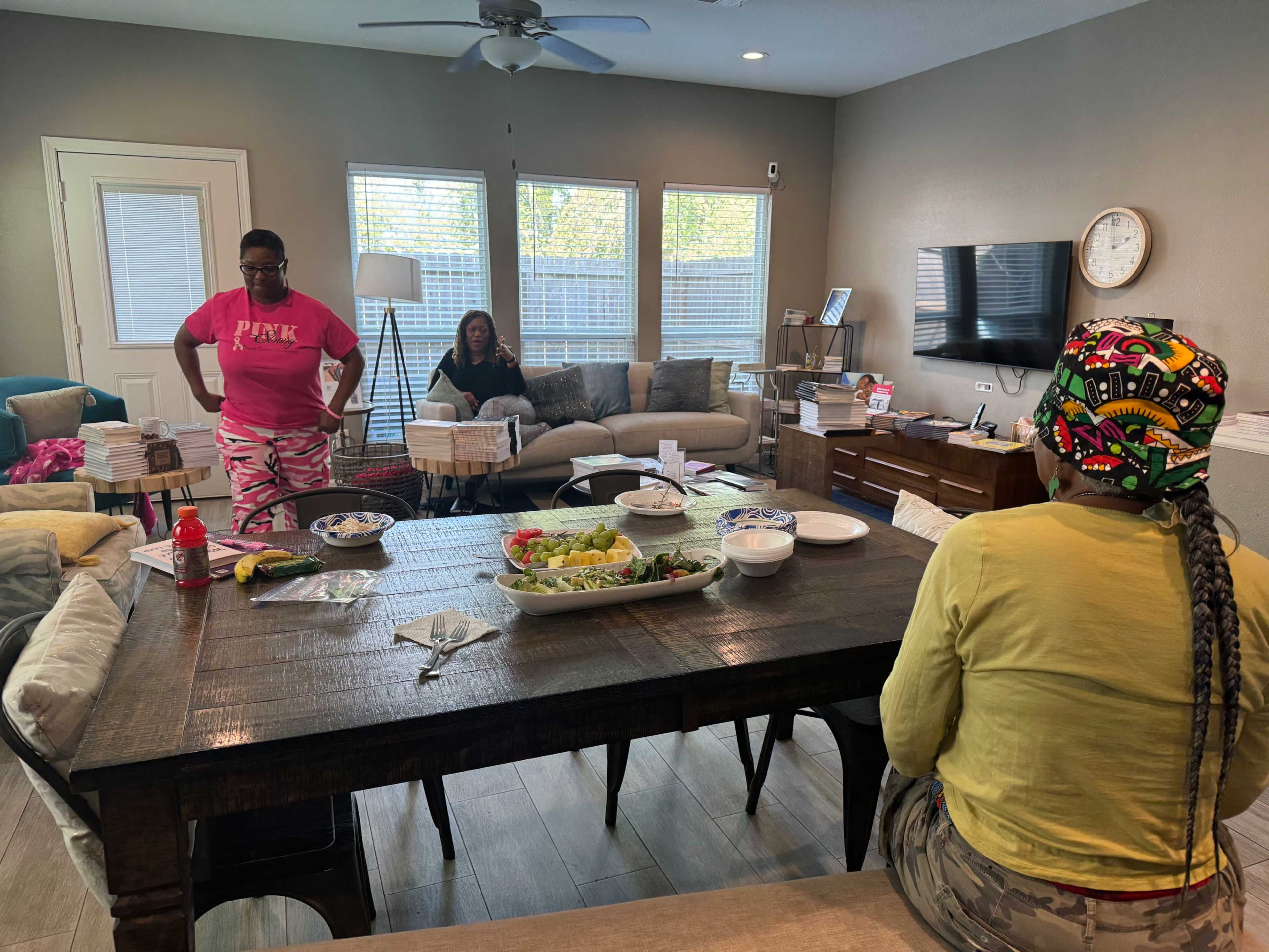 Three women are gathered in a living room, with one person seated at a dining table filled with food, while the others stand nearby.
