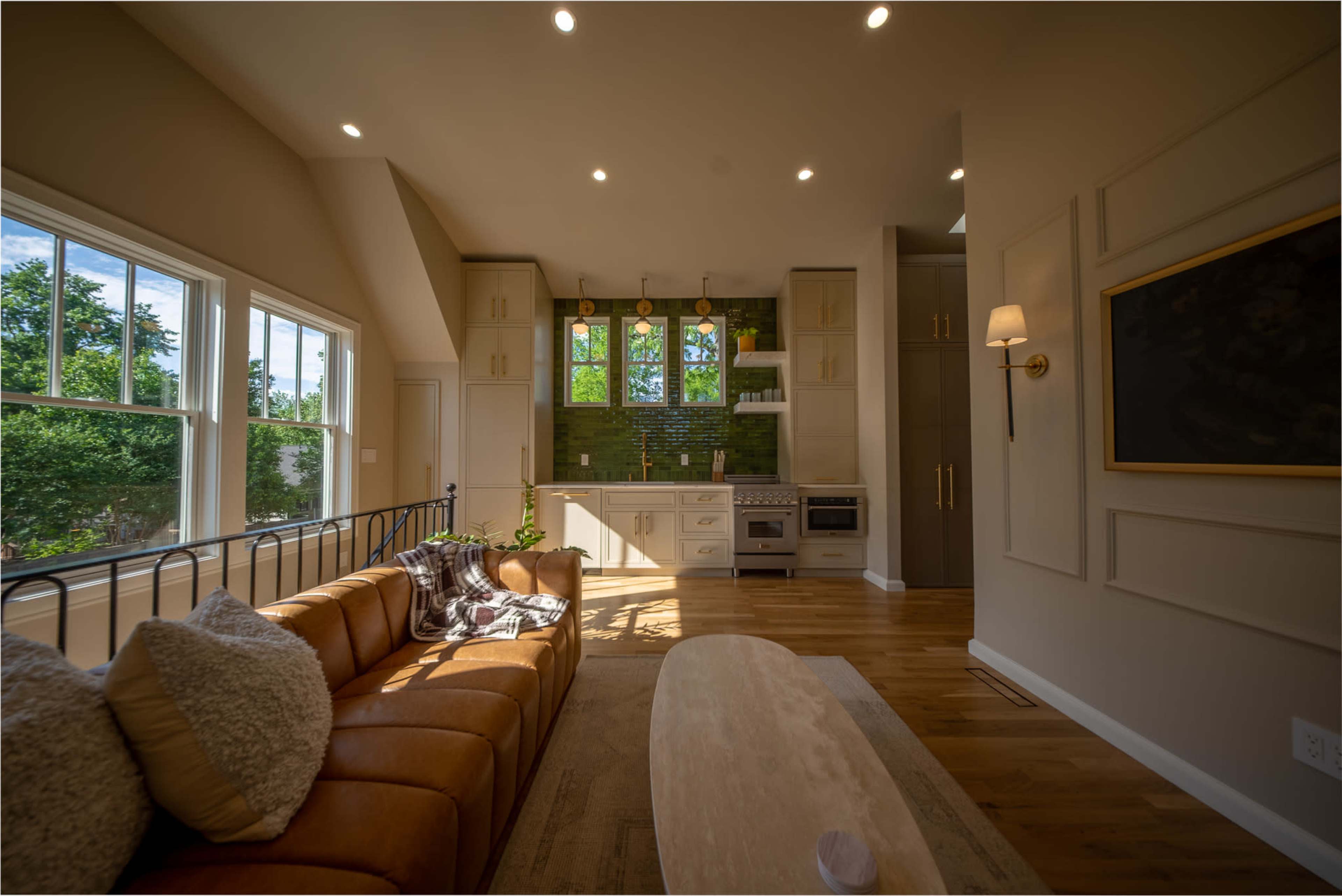 The image shows a modern living room with a brown leather sofa and a kitchen area featuring green tile accents and large windows that let in natural light.