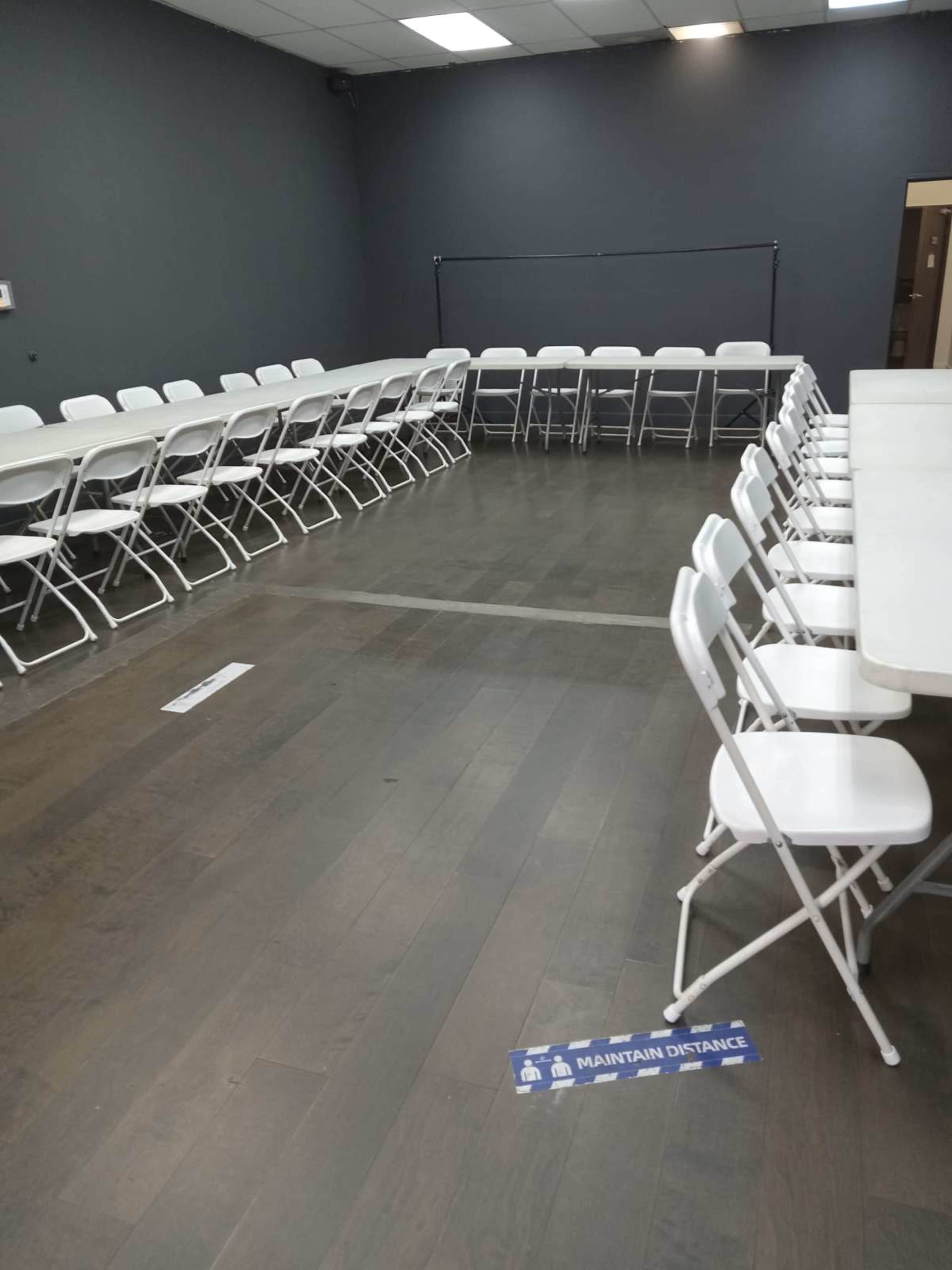 The image shows a spacious room with two rows of folding tables and chairs arranged in a U-shape, along with a distance marker on the floor.