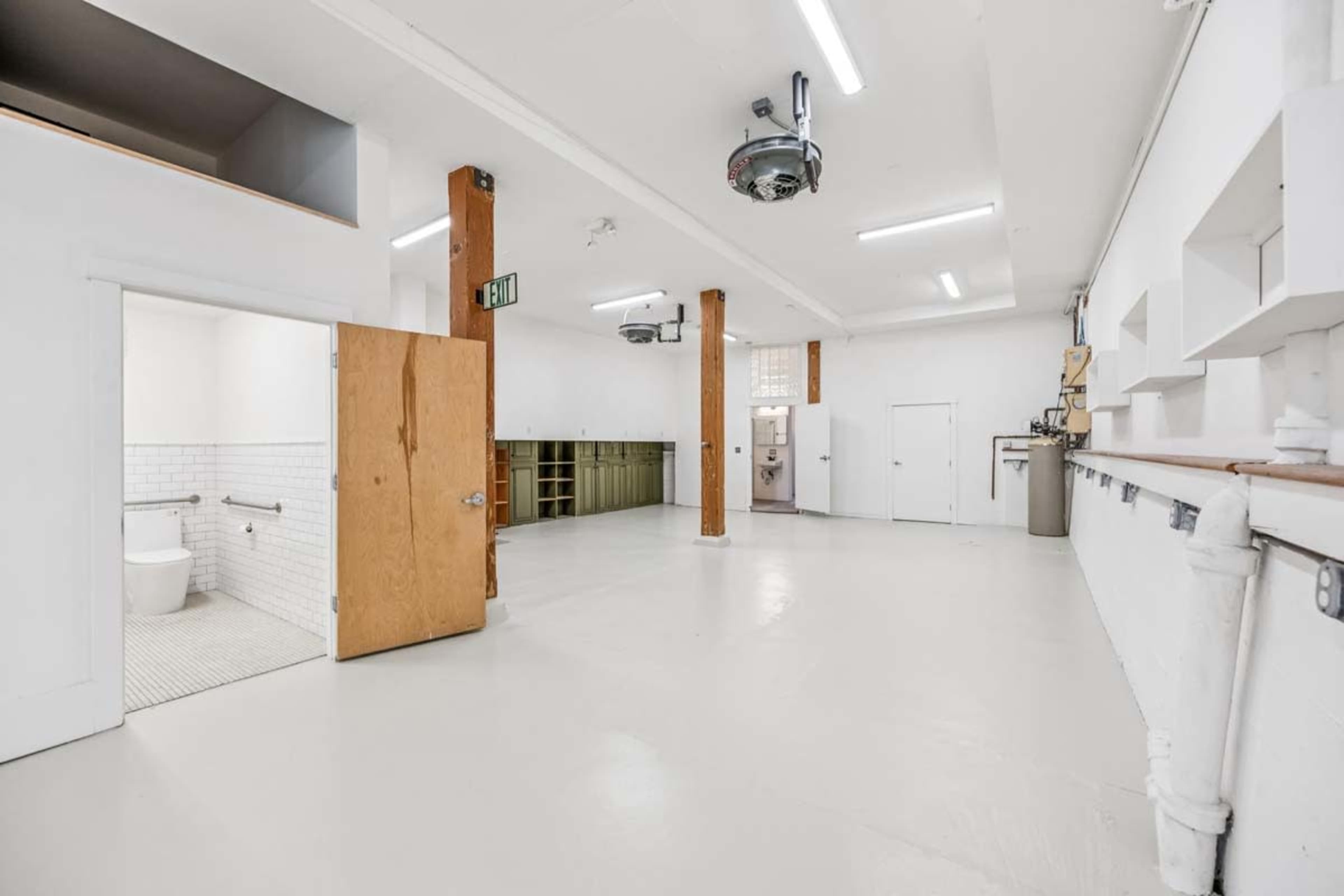 The image shows a spacious, minimalist interior with white walls, a light-colored floor, wooden support columns, and a bathroom area visible in the corner.