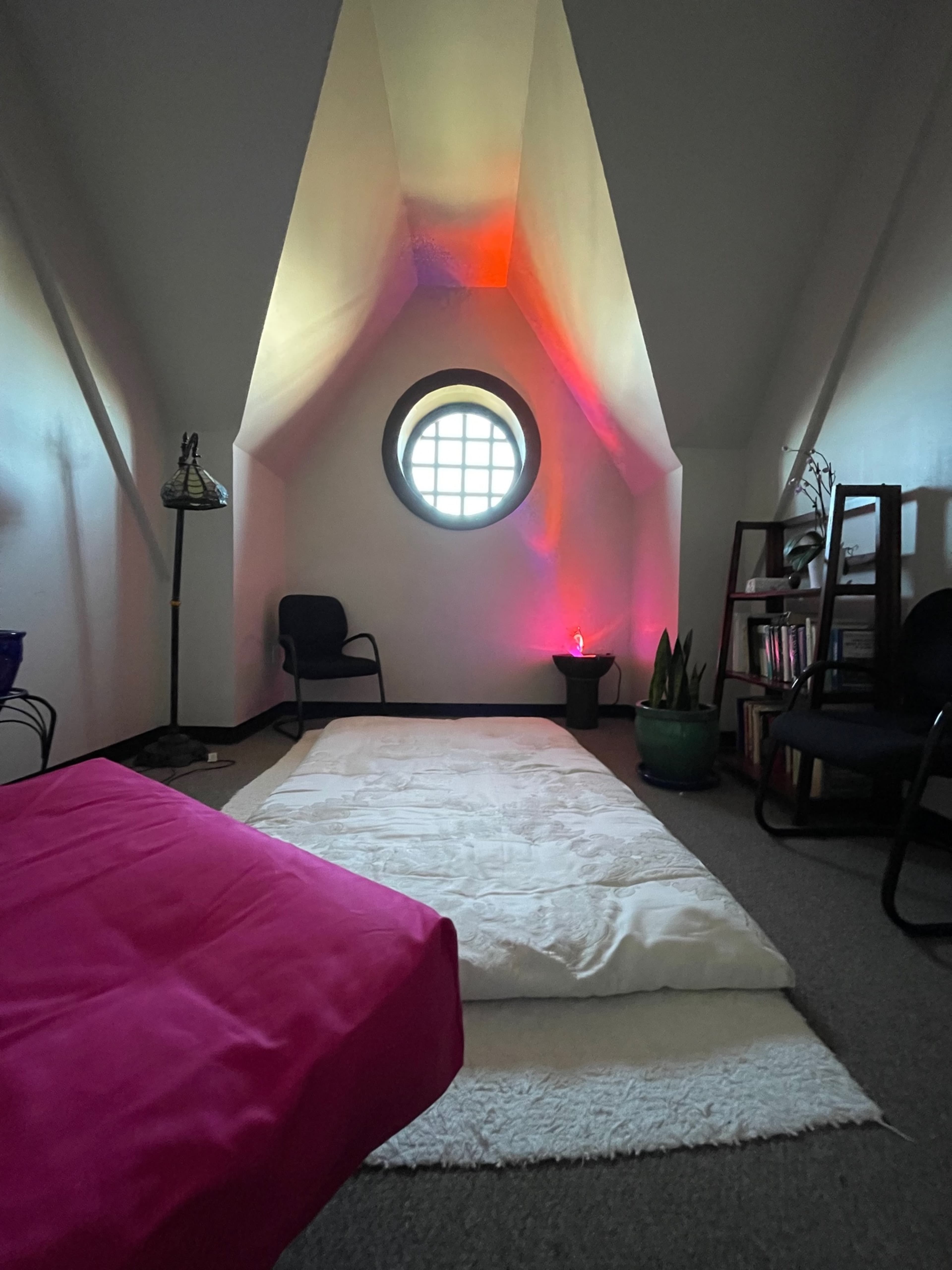 A small, well-lit attic space with a round window, a pink bedspread, a white mattress on the floor, and a bookshelf nearby.