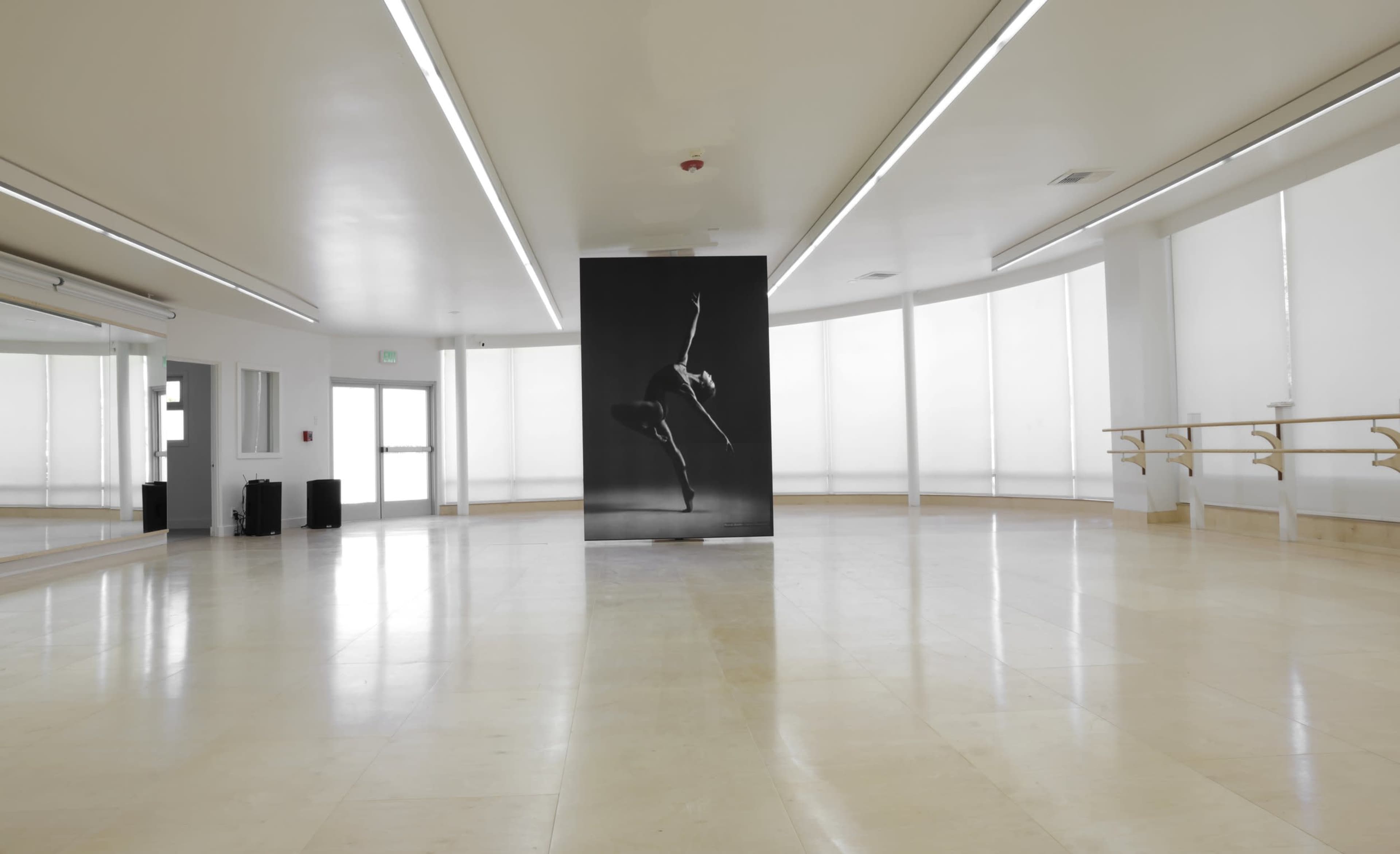 The image shows a spacious ballet studio with large windows, mirrors, a ballet barre, and a prominent black and white photograph of a dancer on display.