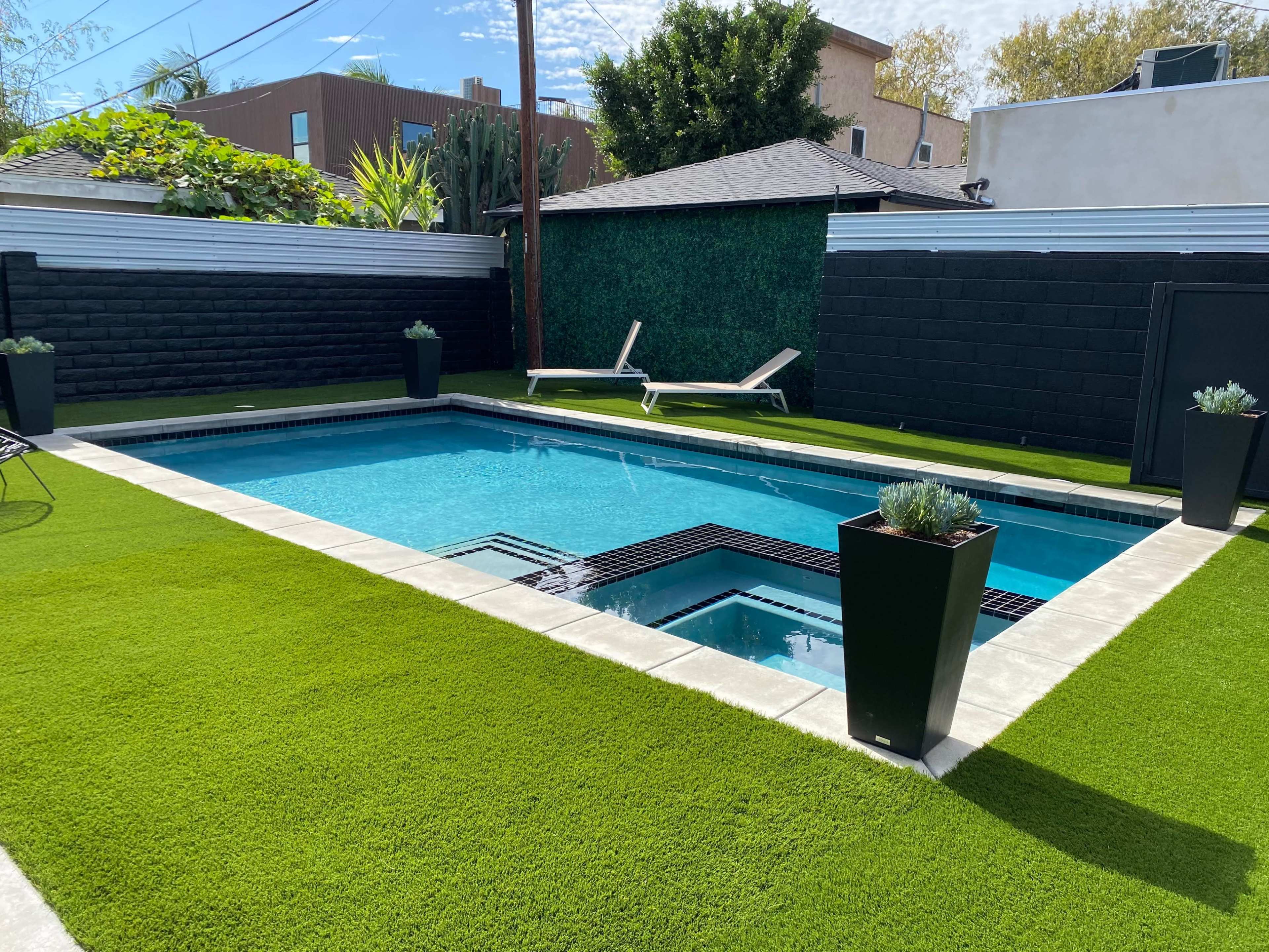 The image shows a rectangular swimming pool surrounded by artificial grass and black walls, with two lounge chairs positioned alongside.