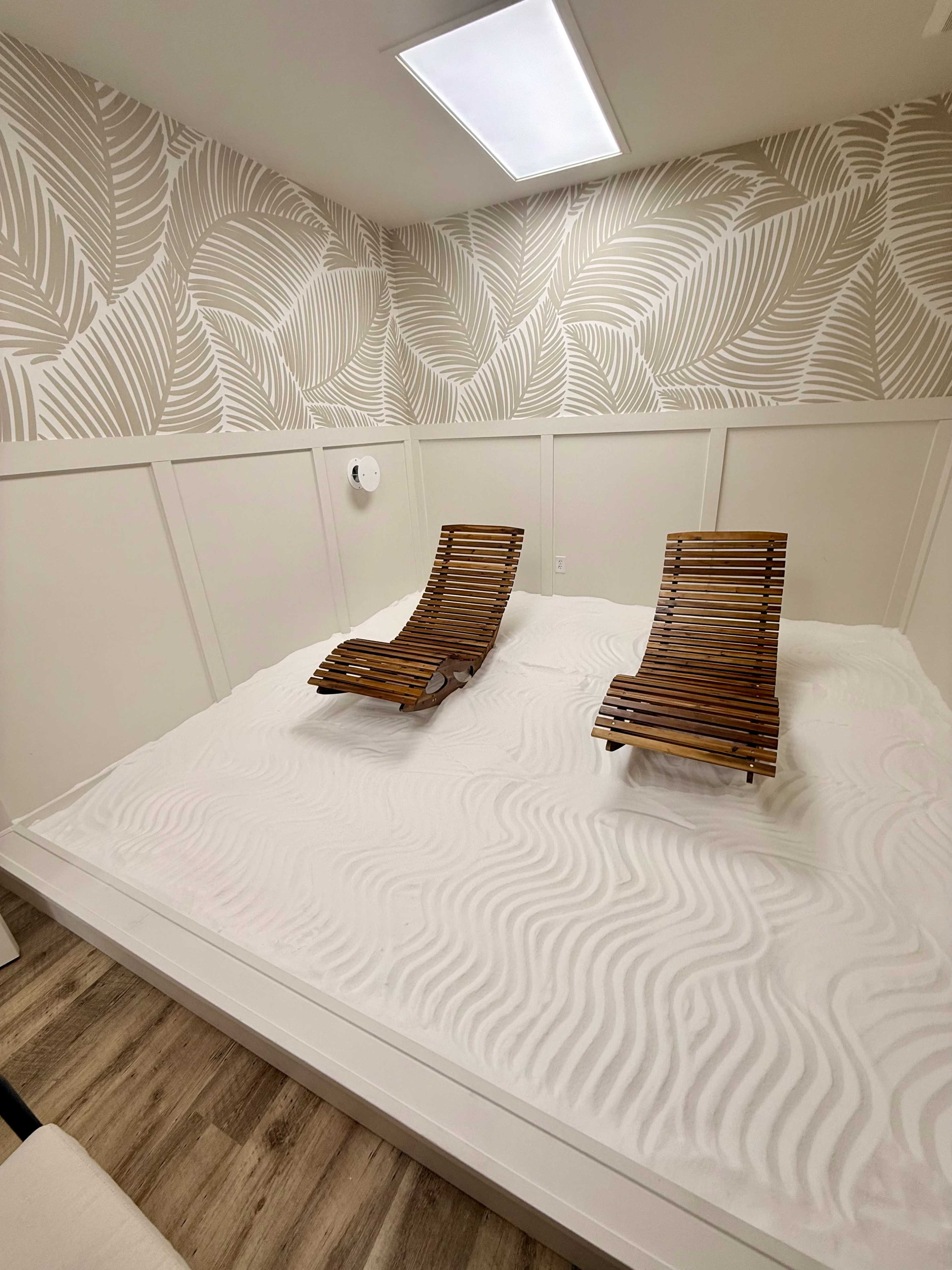 The image shows a small room with two wooden lounge chairs placed on a surface of white sand, surrounded by a wall featuring a leaf-patterned design.