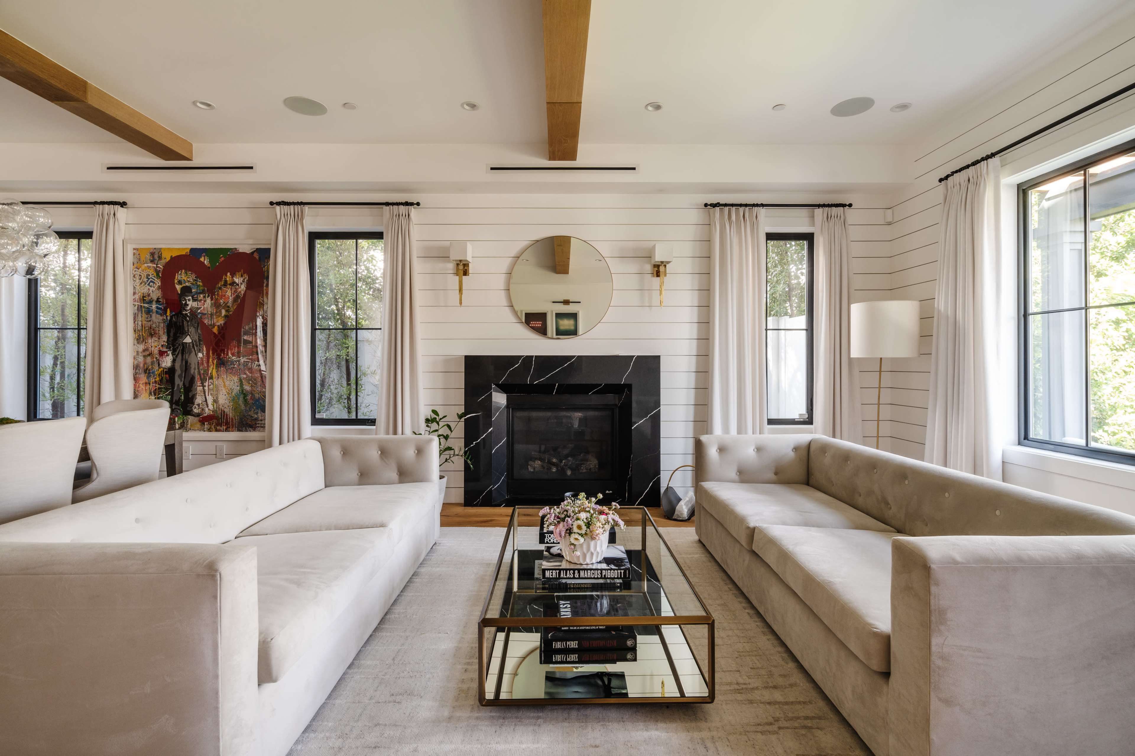 A modern living room with two light-colored sofas facing a glass coffee table in front of a black fireplace, surrounded by large windows and light-colored walls.