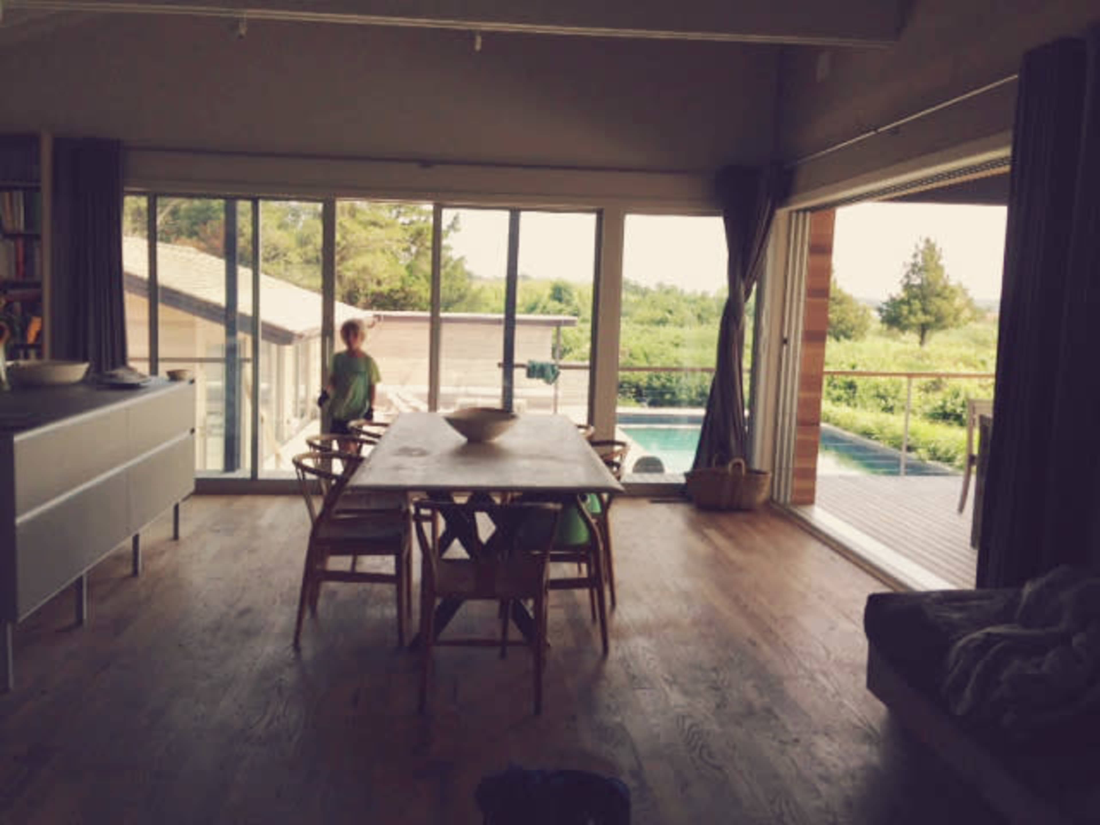 A bright dining area features a wooden table surrounded by chairs, with large windows overlooking a pool and greenery.