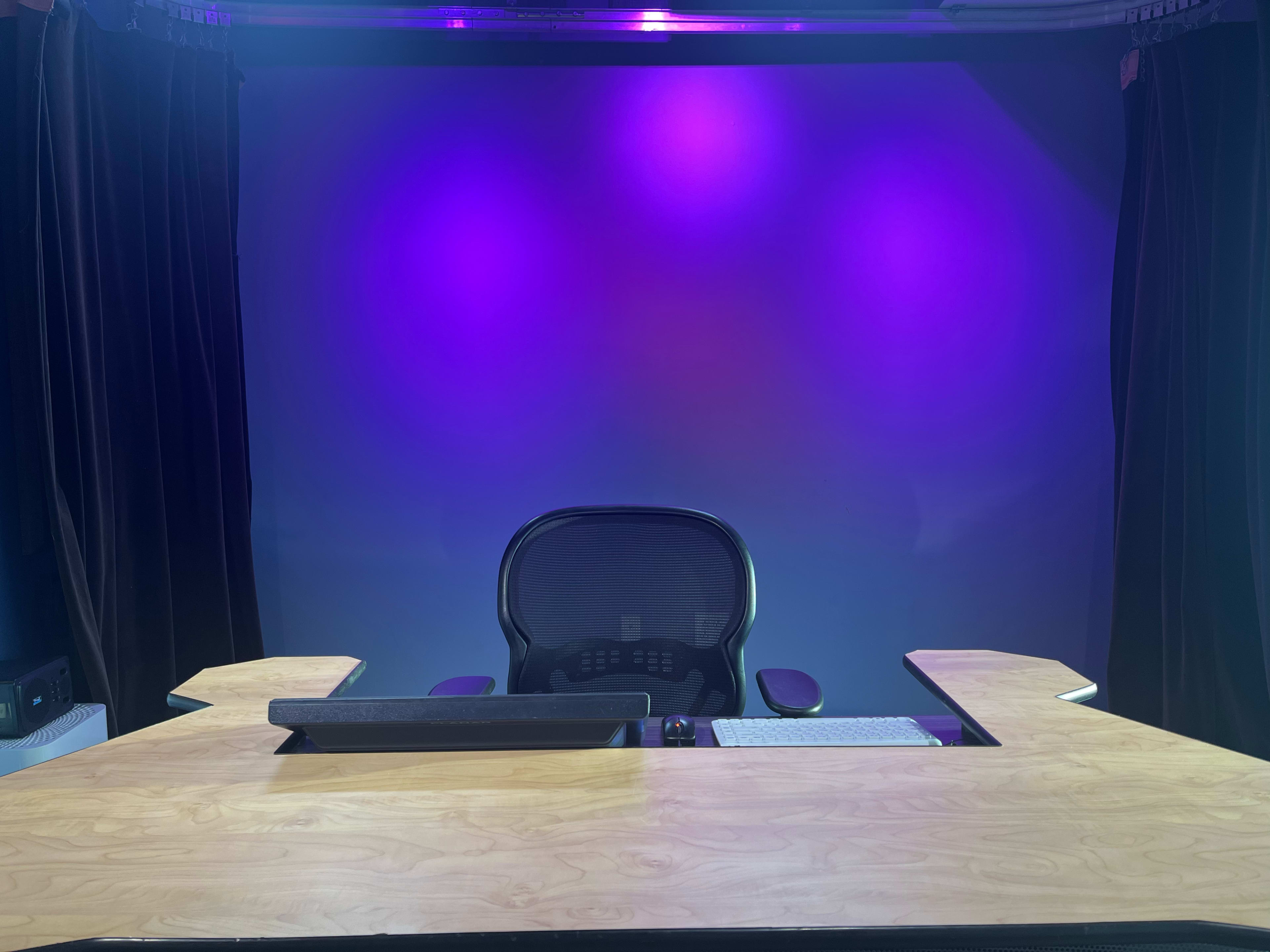 A modern, minimalistic office setup features a wooden desk with a computer monitor and a black ergonomic chair against a backdrop of purple ambient lighting.
