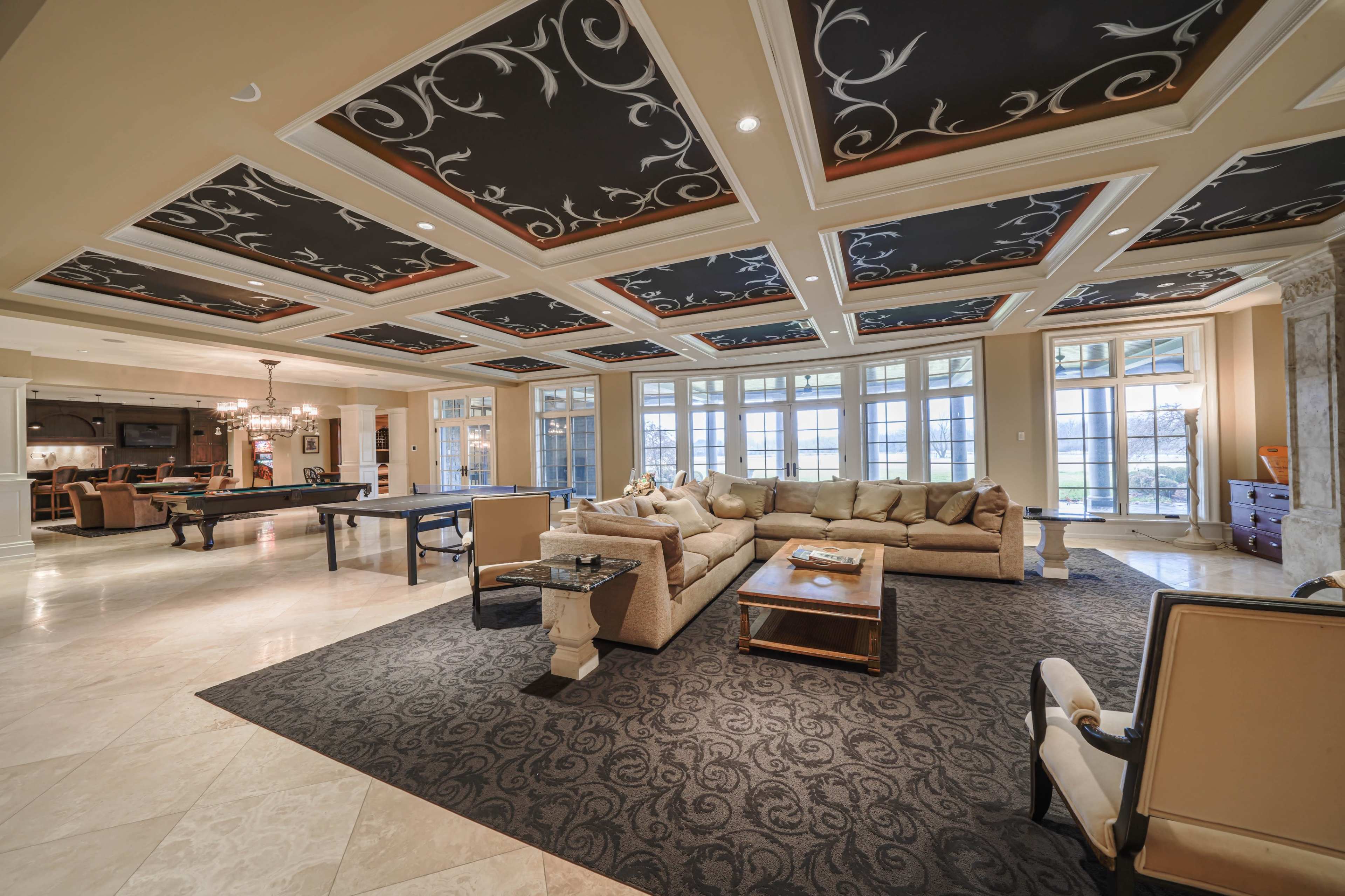 A spacious living room with a dark-patterned ceiling, large windows, and a seating area featuring a beige sectional sofa, wooden coffee table, and a pool table.