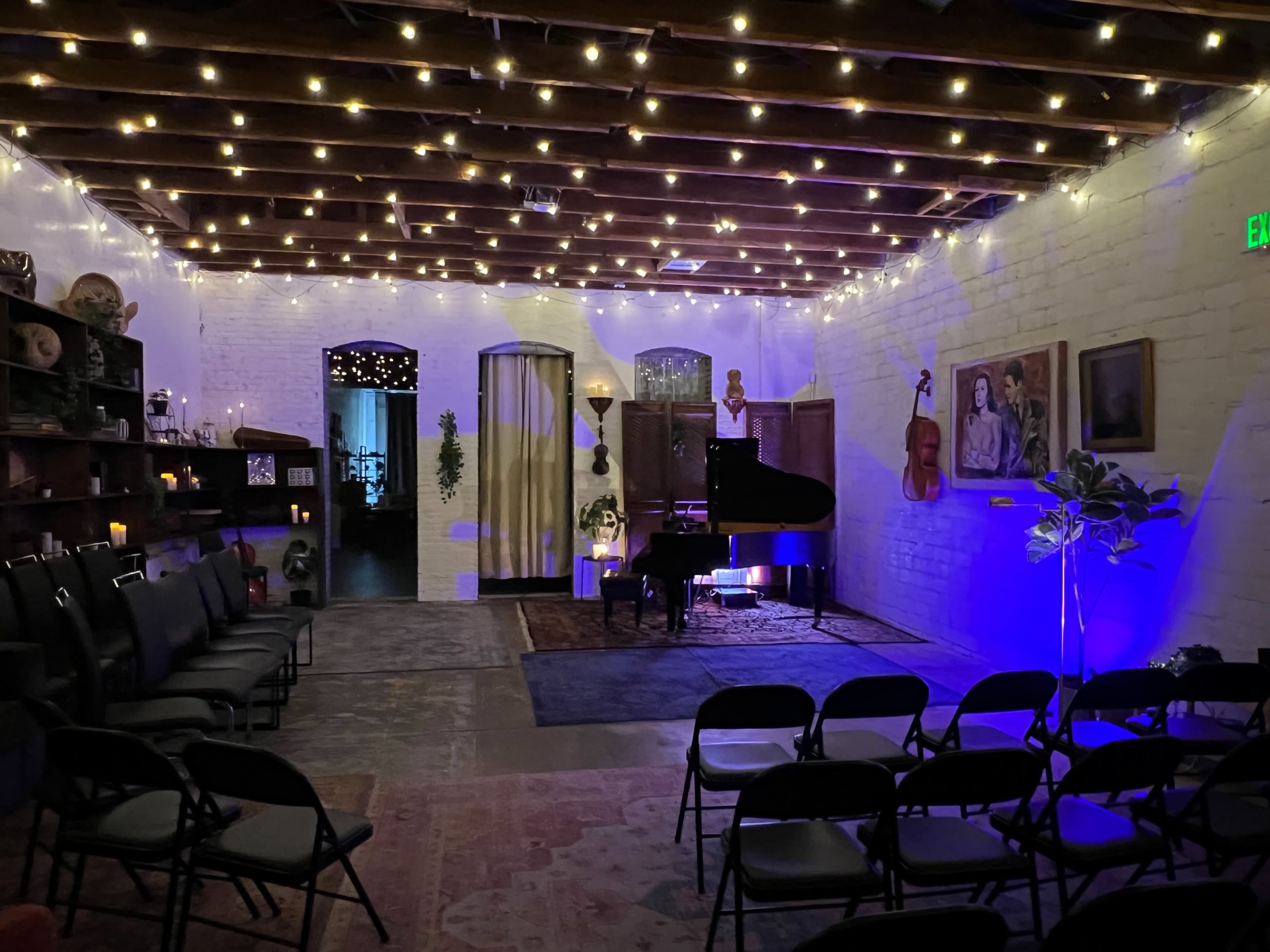 The image shows a spacious room with string lights overhead, a grand piano, and arranged chairs facing the performance area, creating a setting for a gathering or event.