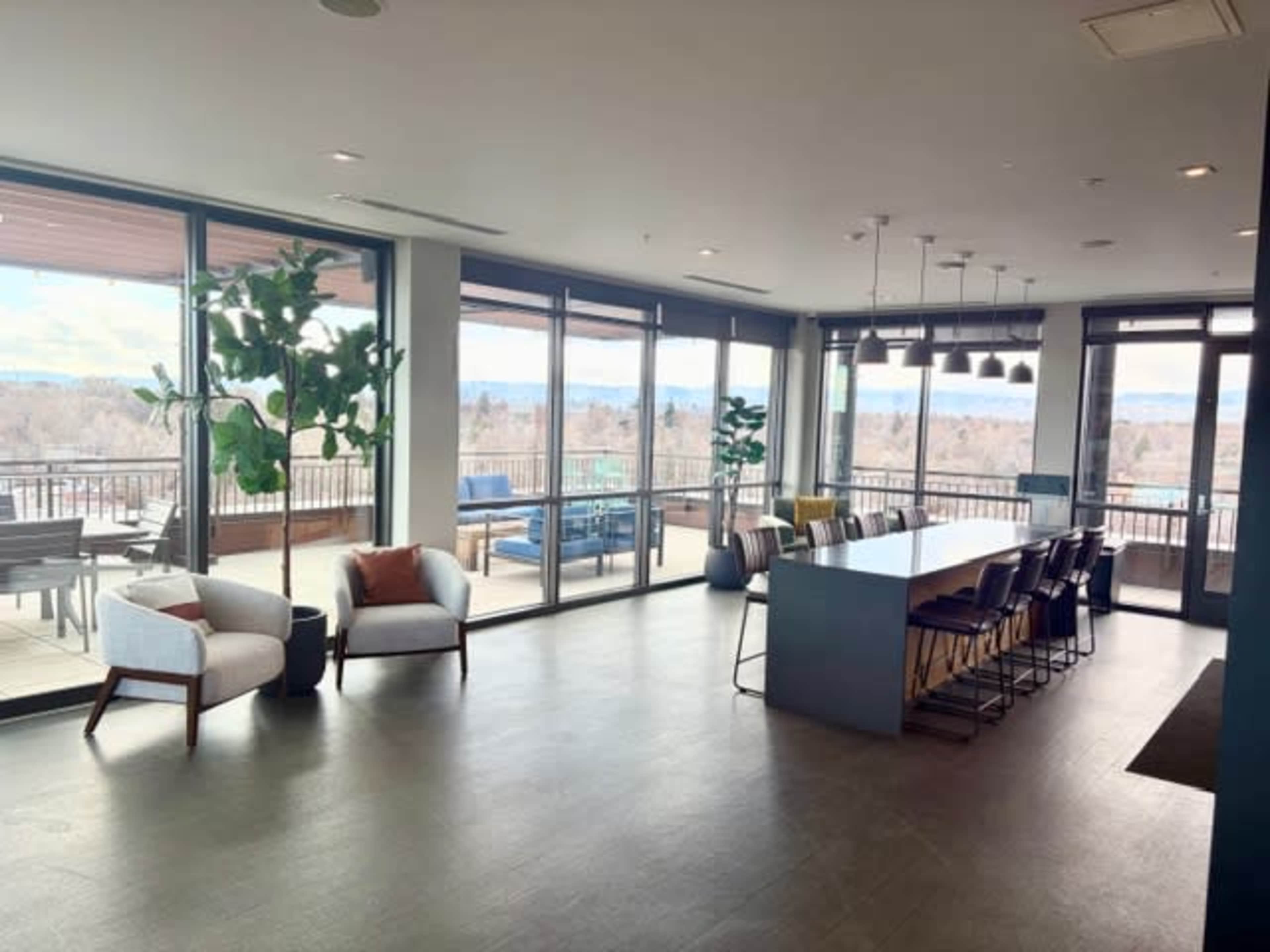 The image shows a modern, spacious interior lounge area with large windows, plants, comfortable seating, and a long dining table.