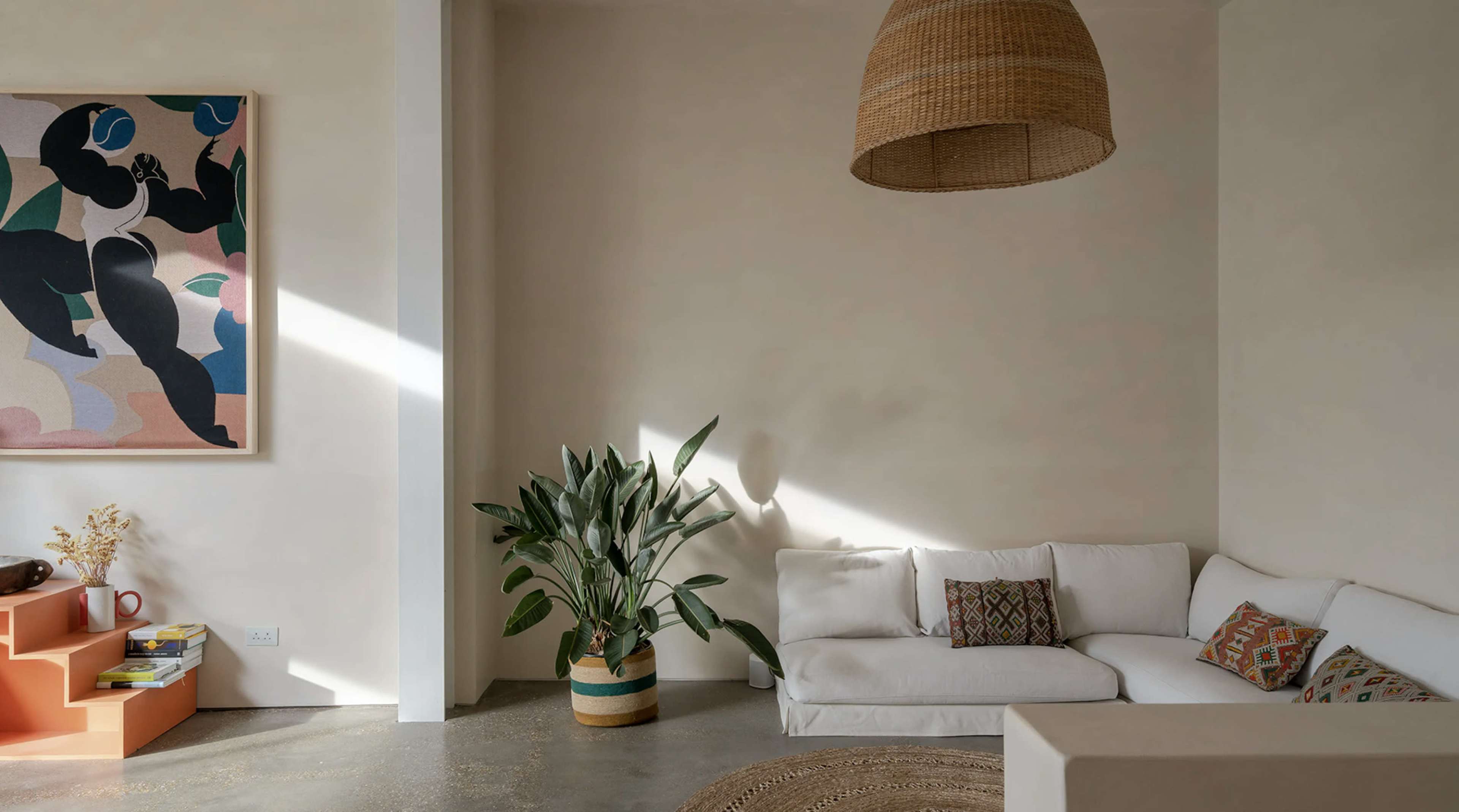 A light-colored room with a white sofa, a potted plant, and a large artwork on the wall, illuminated by natural light.