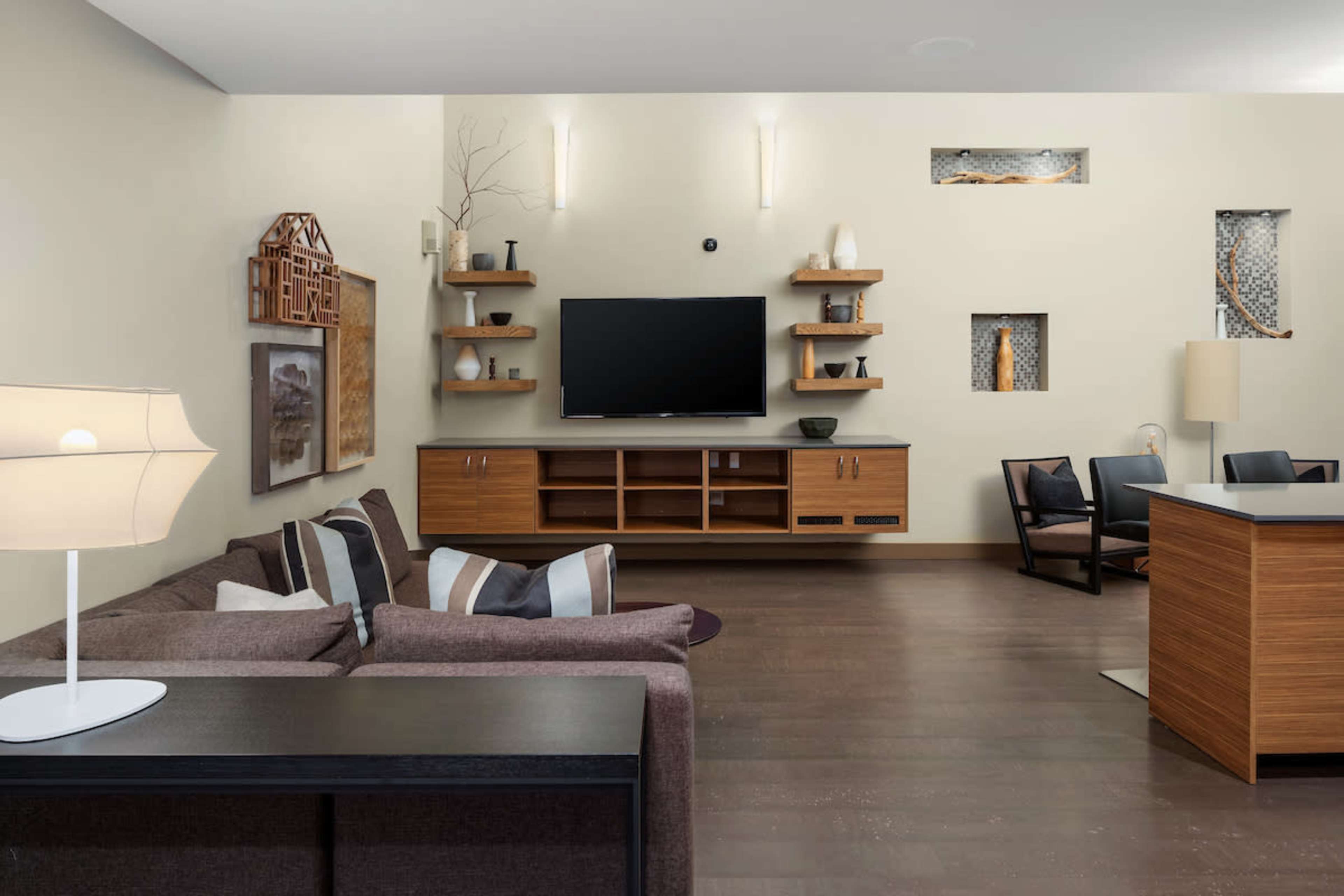 A modern living room with a brown sectional sofa, a flat-screen TV mounted on a light-colored wall, wooden shelving with decorative items, and a dining area with a black table and chairs.