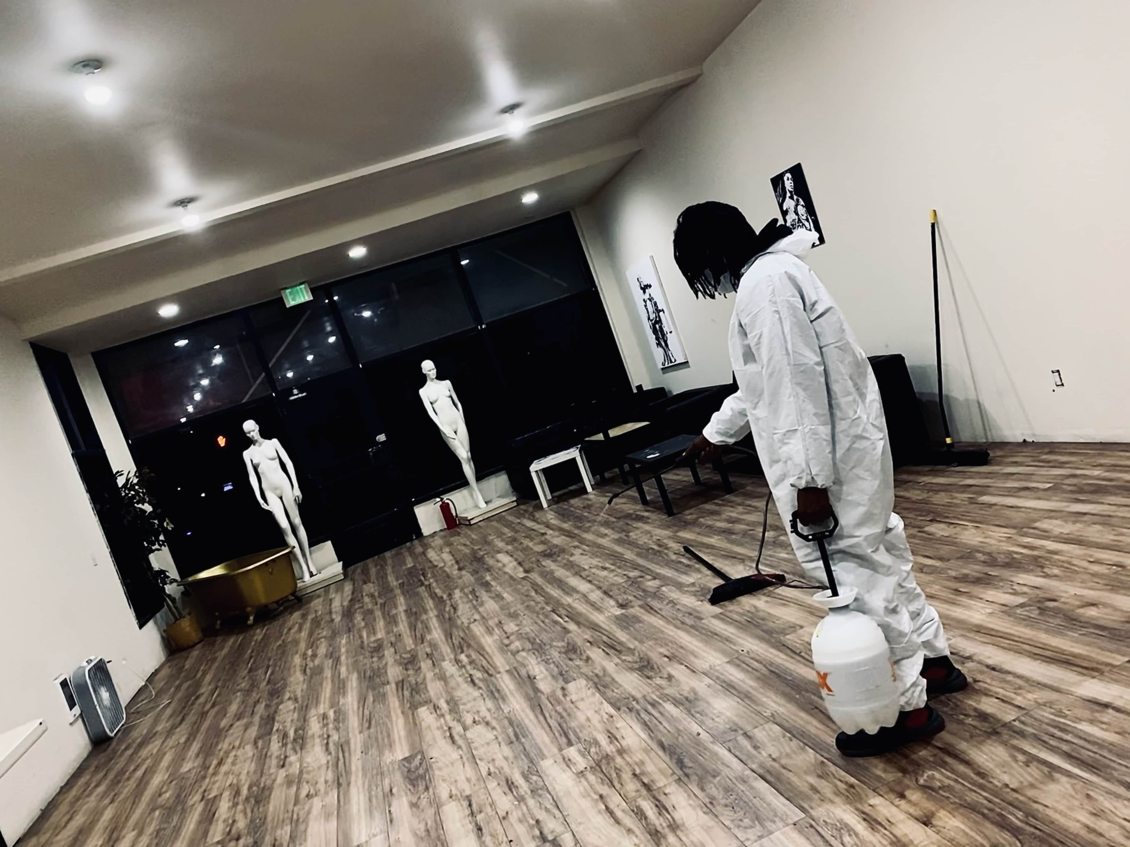 A person in a protective suit is using a sprayer in a spacious room with mannequins positioned near large windows.