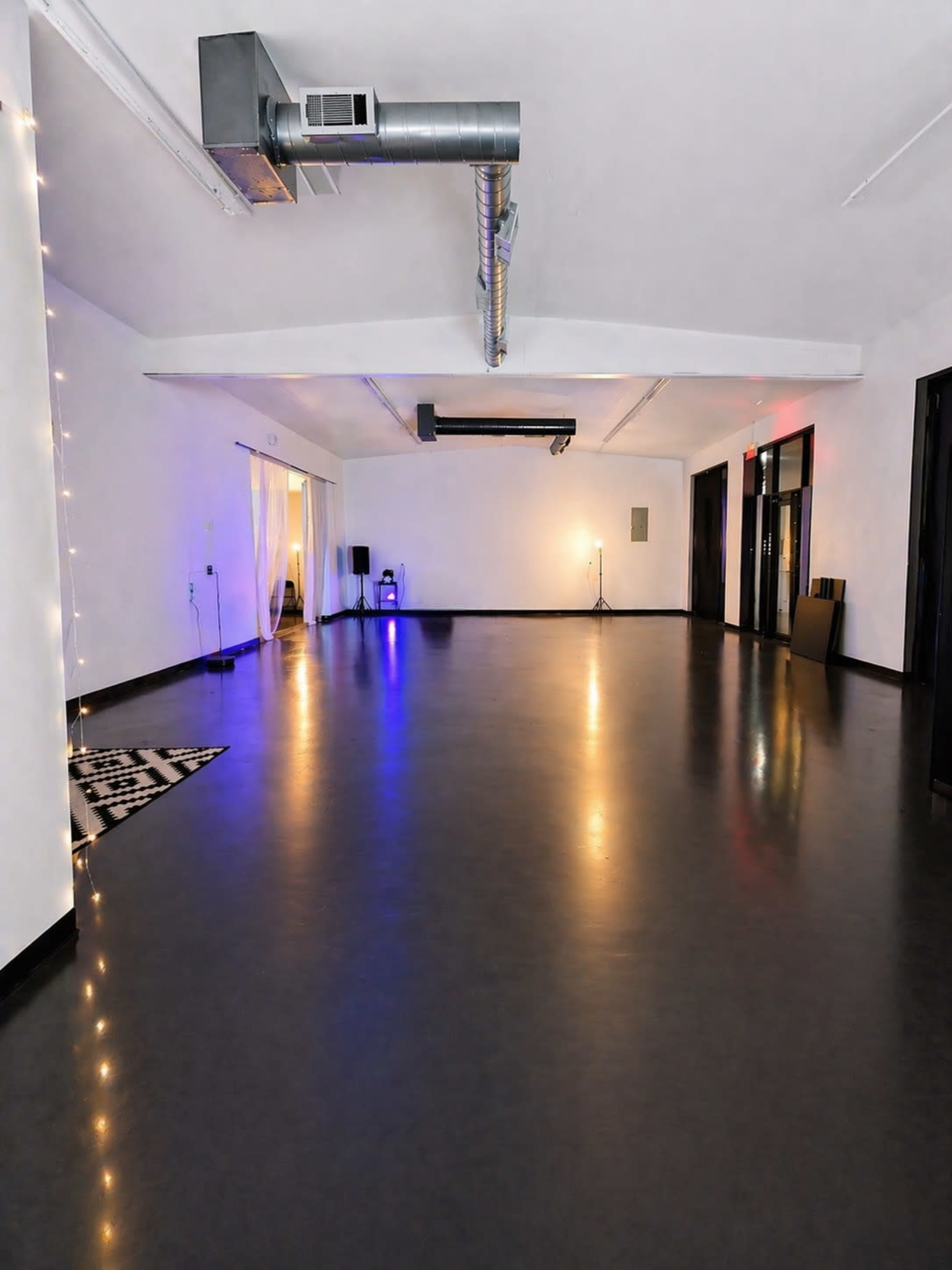 The image shows a spacious, empty room with polished dark flooring, dim lighting, and minimal decor, featuring a few light sources and a carpet.