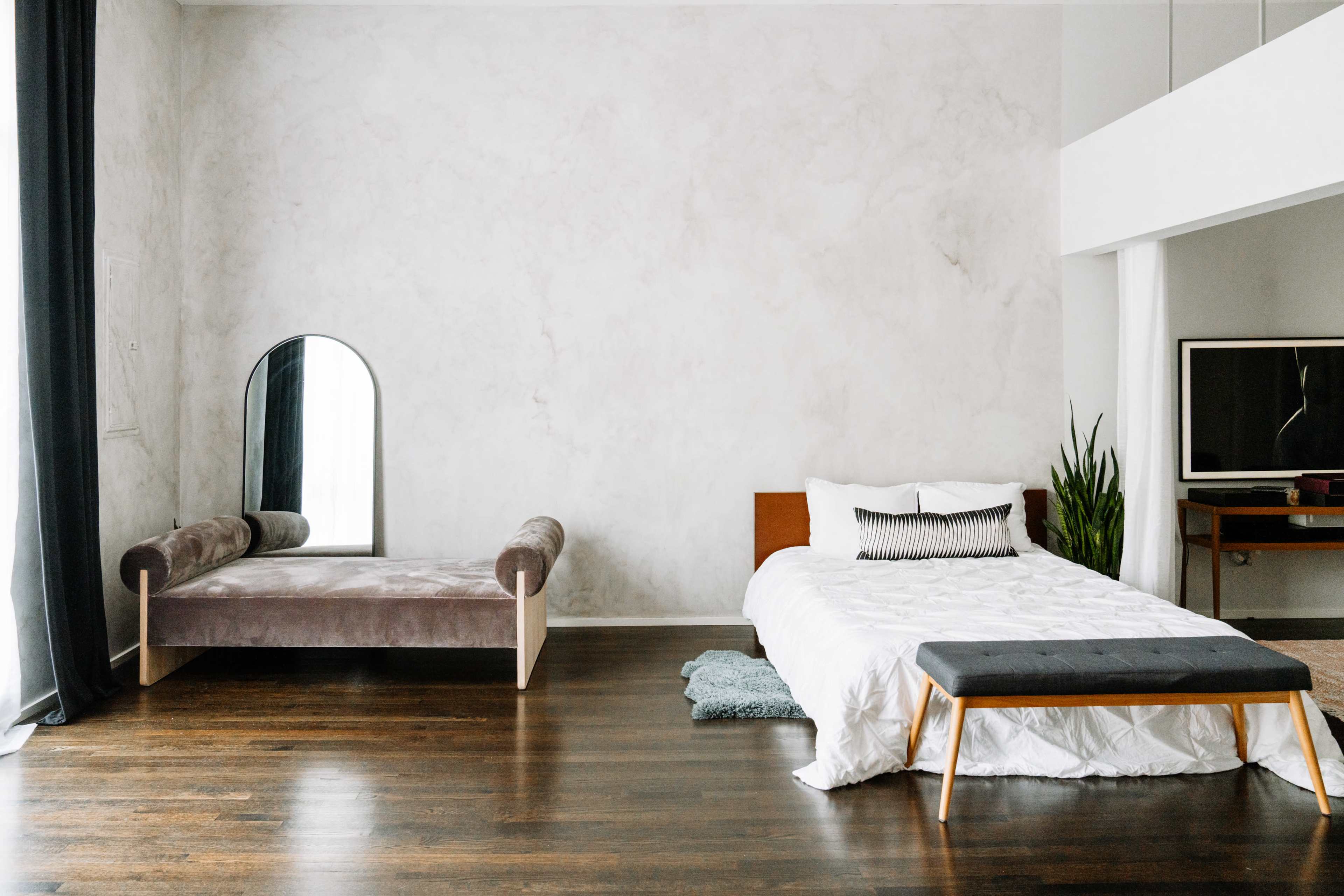 A minimalist bedroom with a bed, a bench, a mirror, a plant, and a television on a wooden floor.