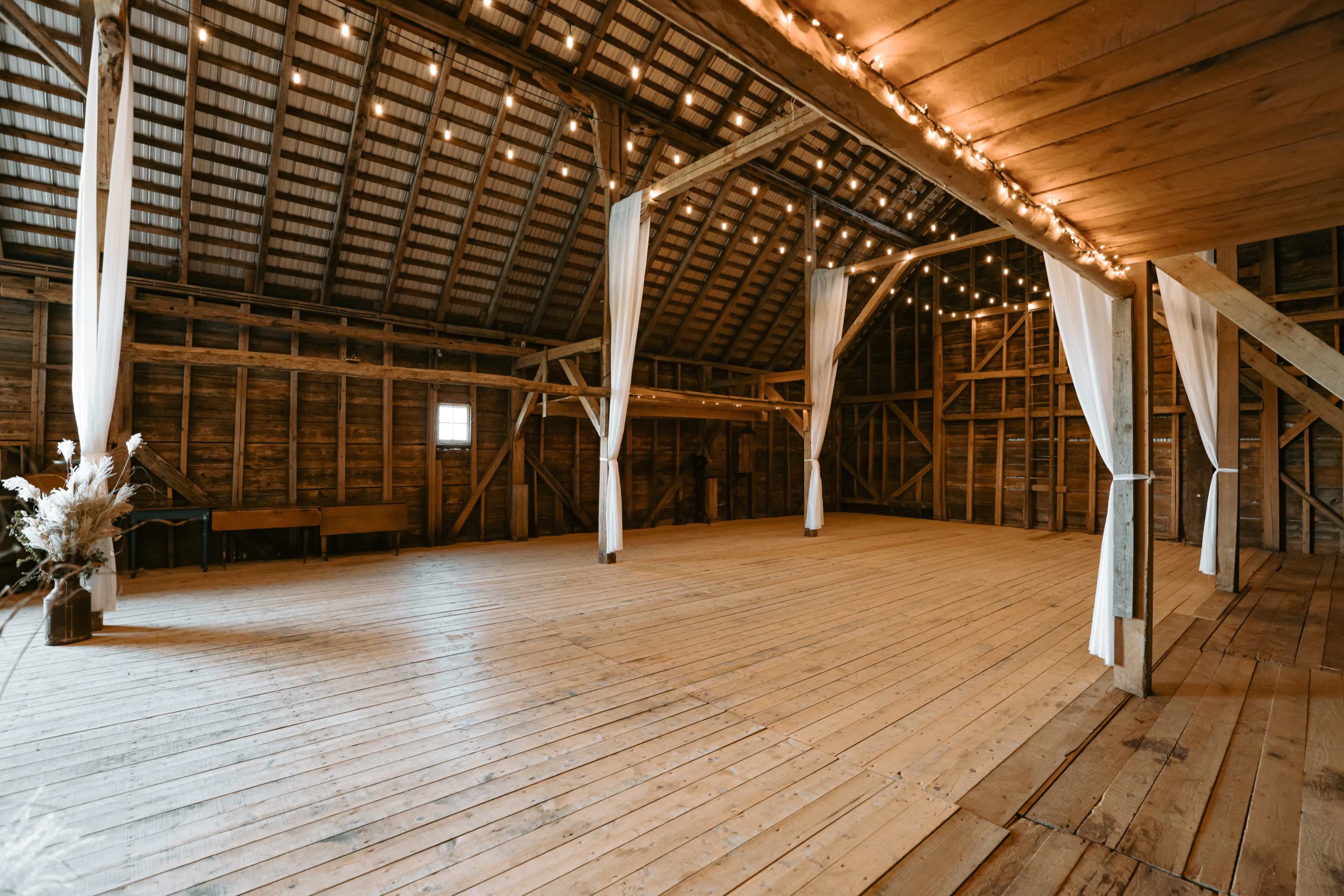 The image shows the interior of a spacious wooden barn with high ceilings, exposed beams, and string lights illuminating the wooden floor.