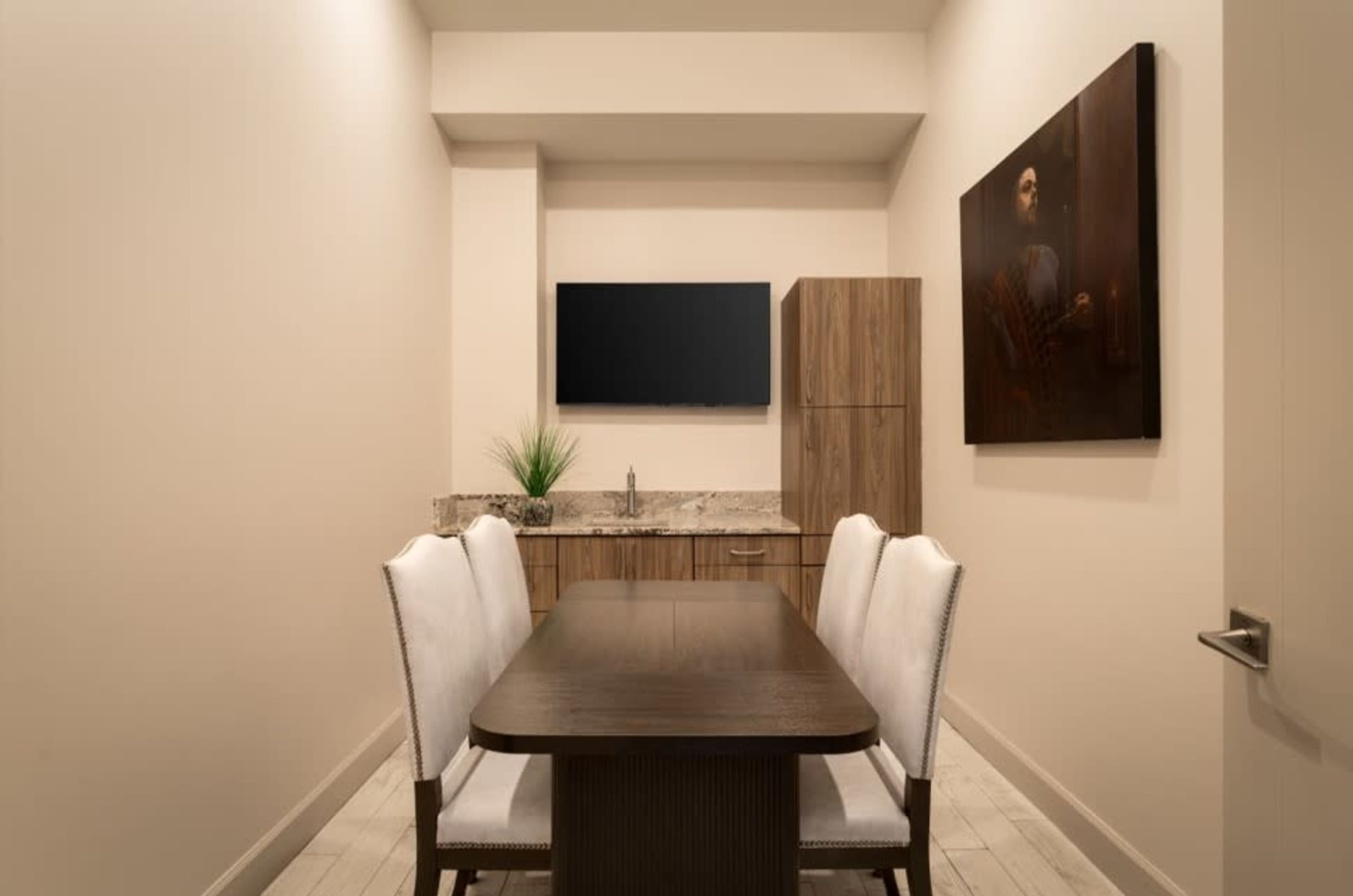 A small meeting room features a wooden table surrounded by four white chairs, with a wall-mounted television and a cabinet in the background.