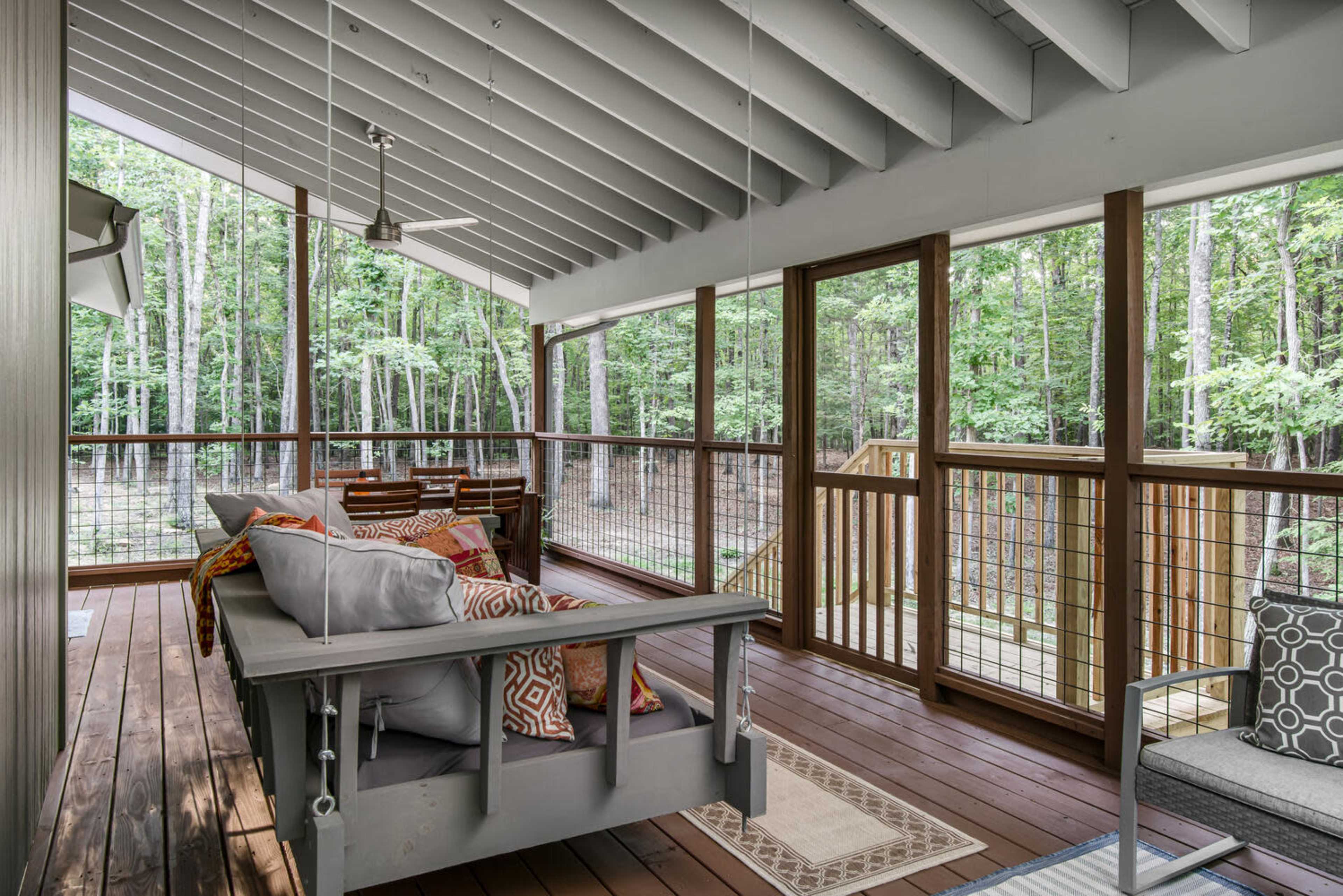 The image shows a wooden porch with a swing and seating area, surrounded by trees in a forested setting.