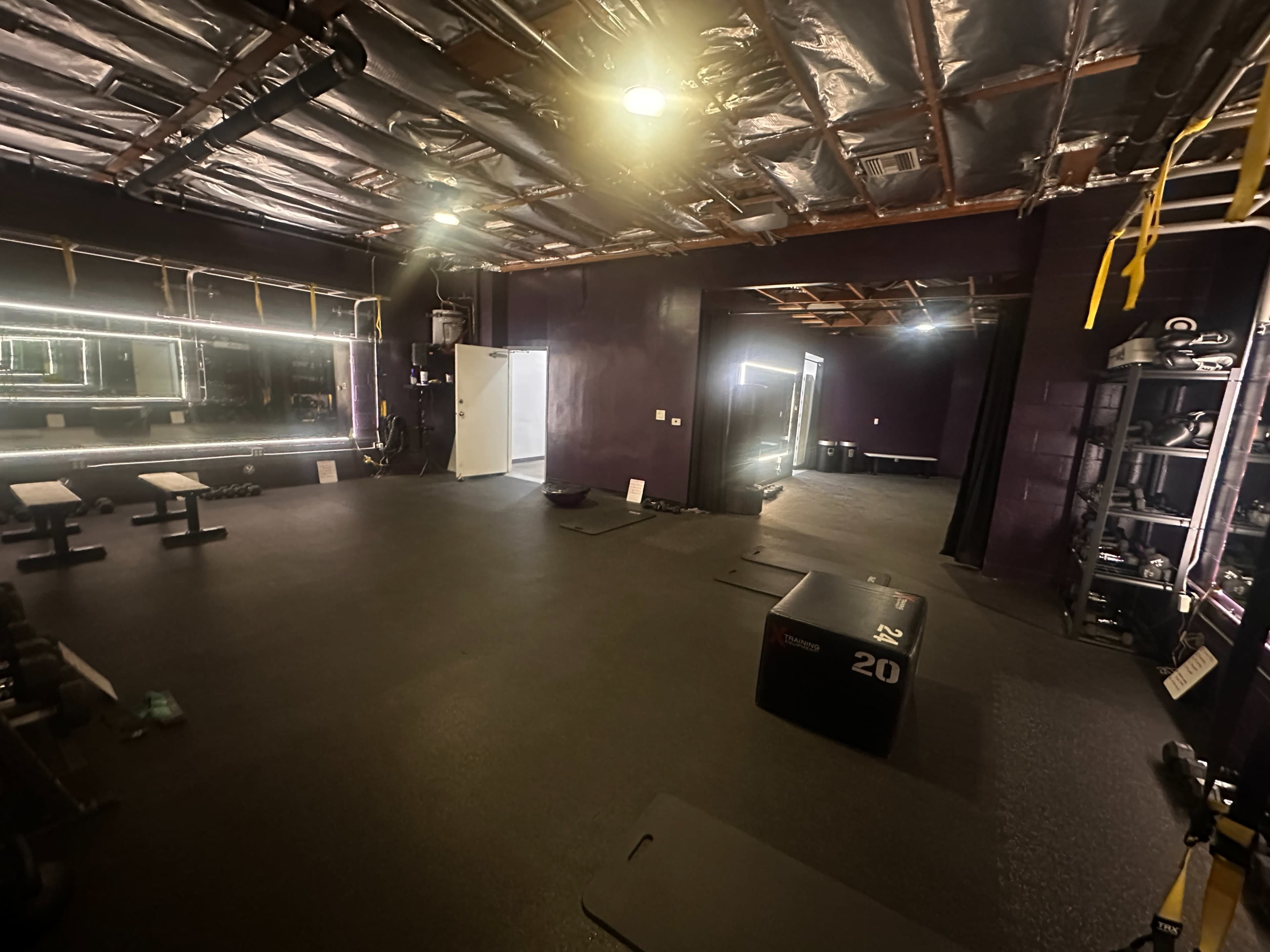 The image shows a dimly lit gym with a mirrored wall, workout benches, dumbbells, and a plyometric box on the floor.