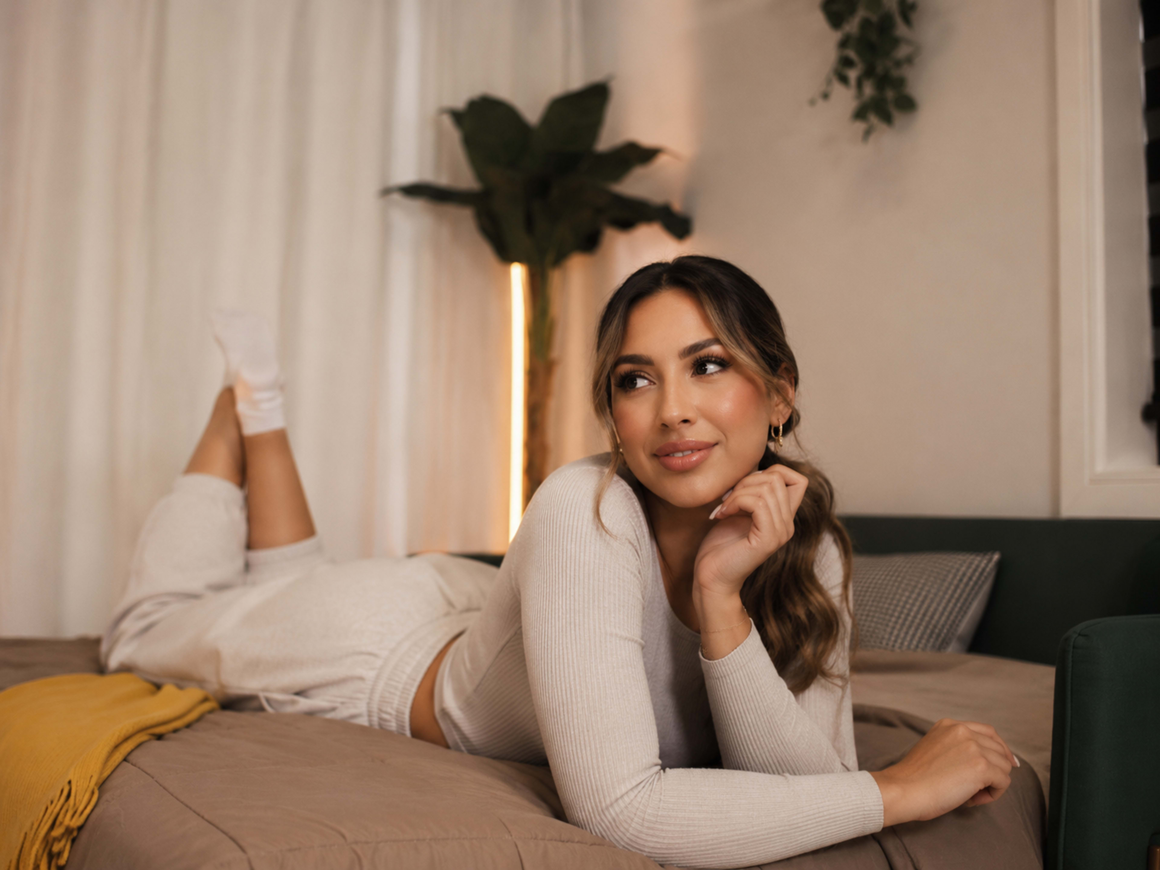 A woman in a cozy setting lies on a bed, resting her chin on her hand while looking thoughtfully to the side.