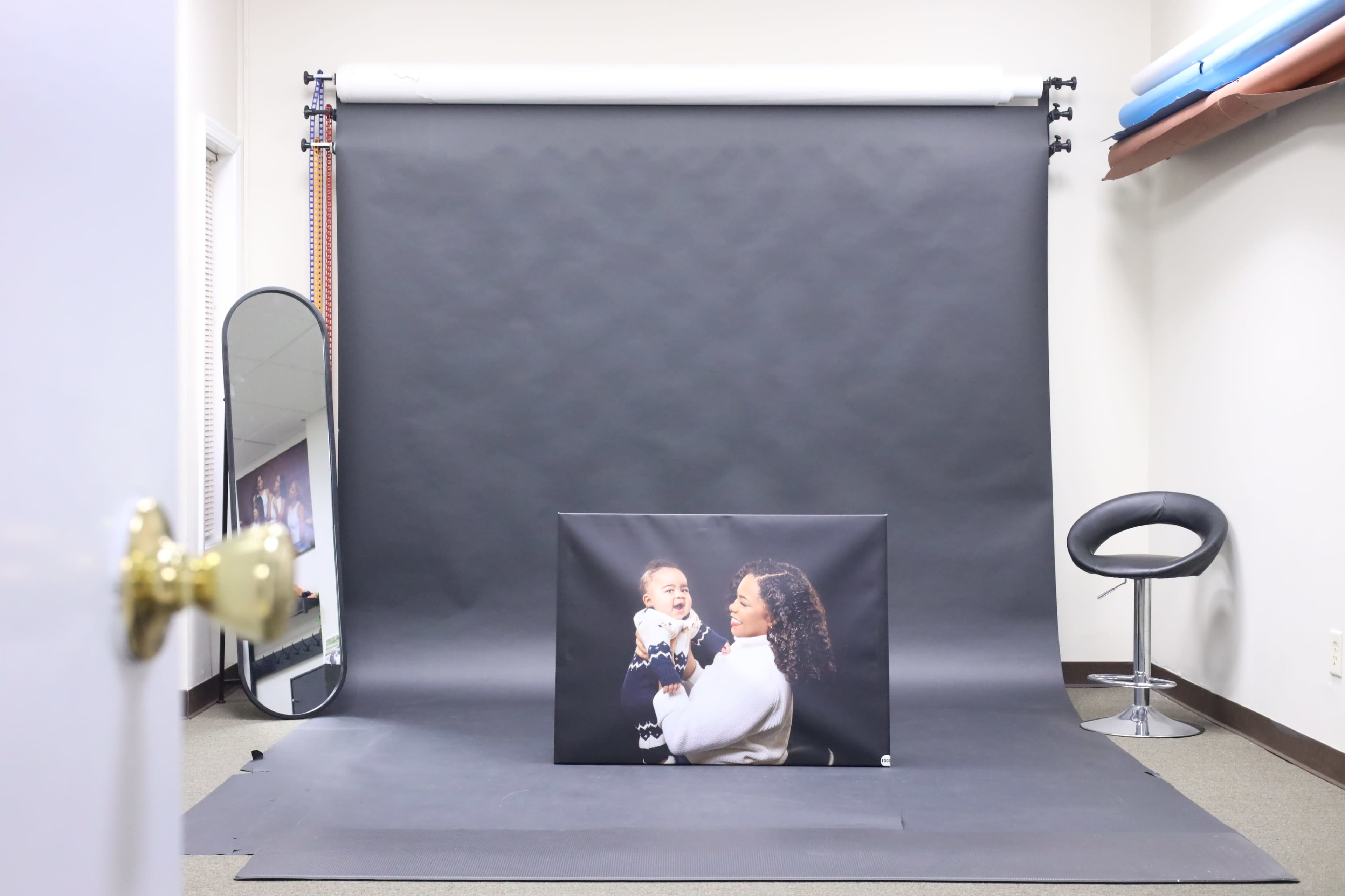 A photography studio is setup with a black backdrop, a large portrait of a woman holding a baby, a mirror, and a chair.
