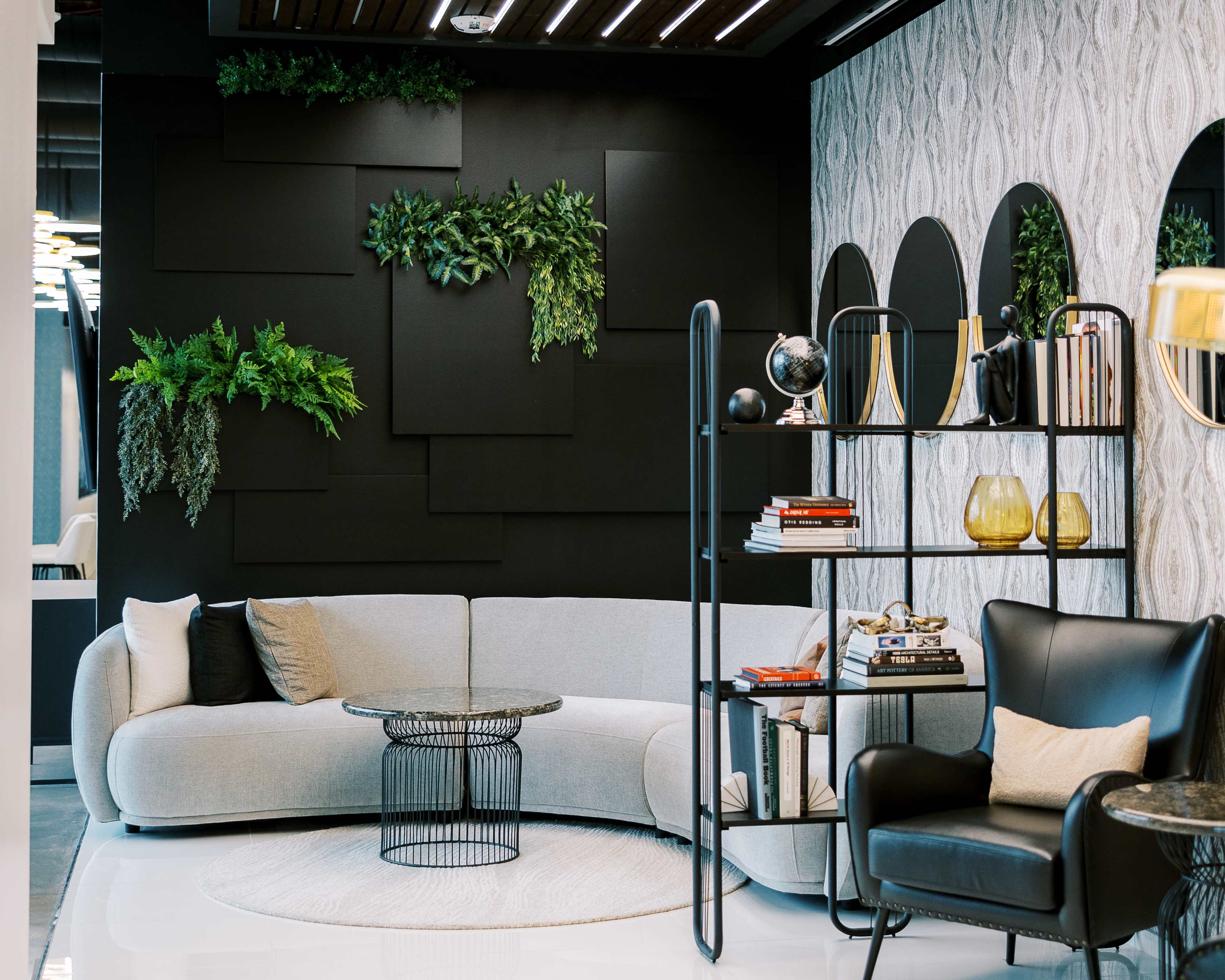 The image shows a modern living room with a curved grey sofa, a circular coffee table, a black shelving unit filled with books and decor, and greenery on the walls.