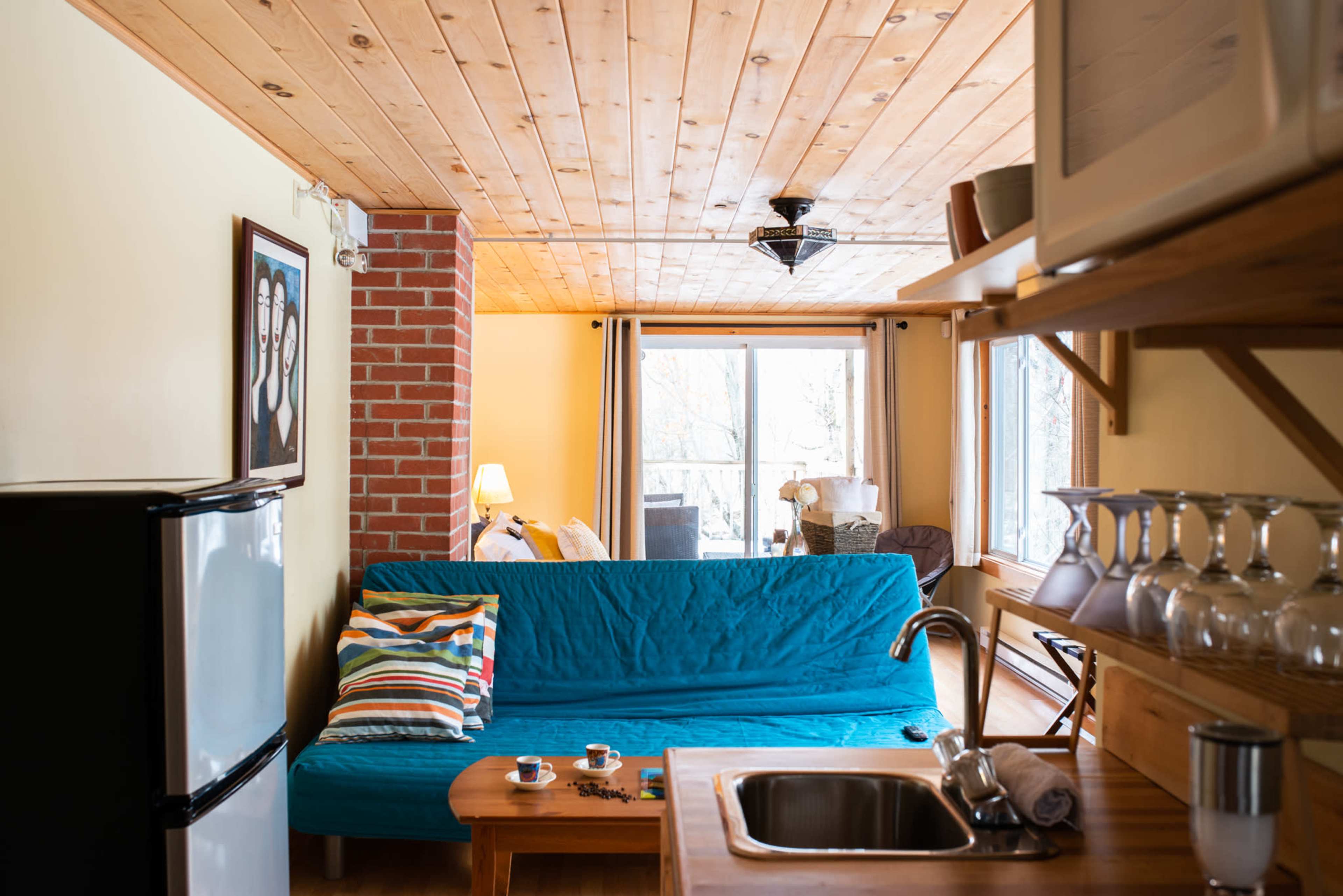 A small living space features a blue sofa, a wooden table with cups, and a kitchenette, with a window allowing natural light to illuminate the room.