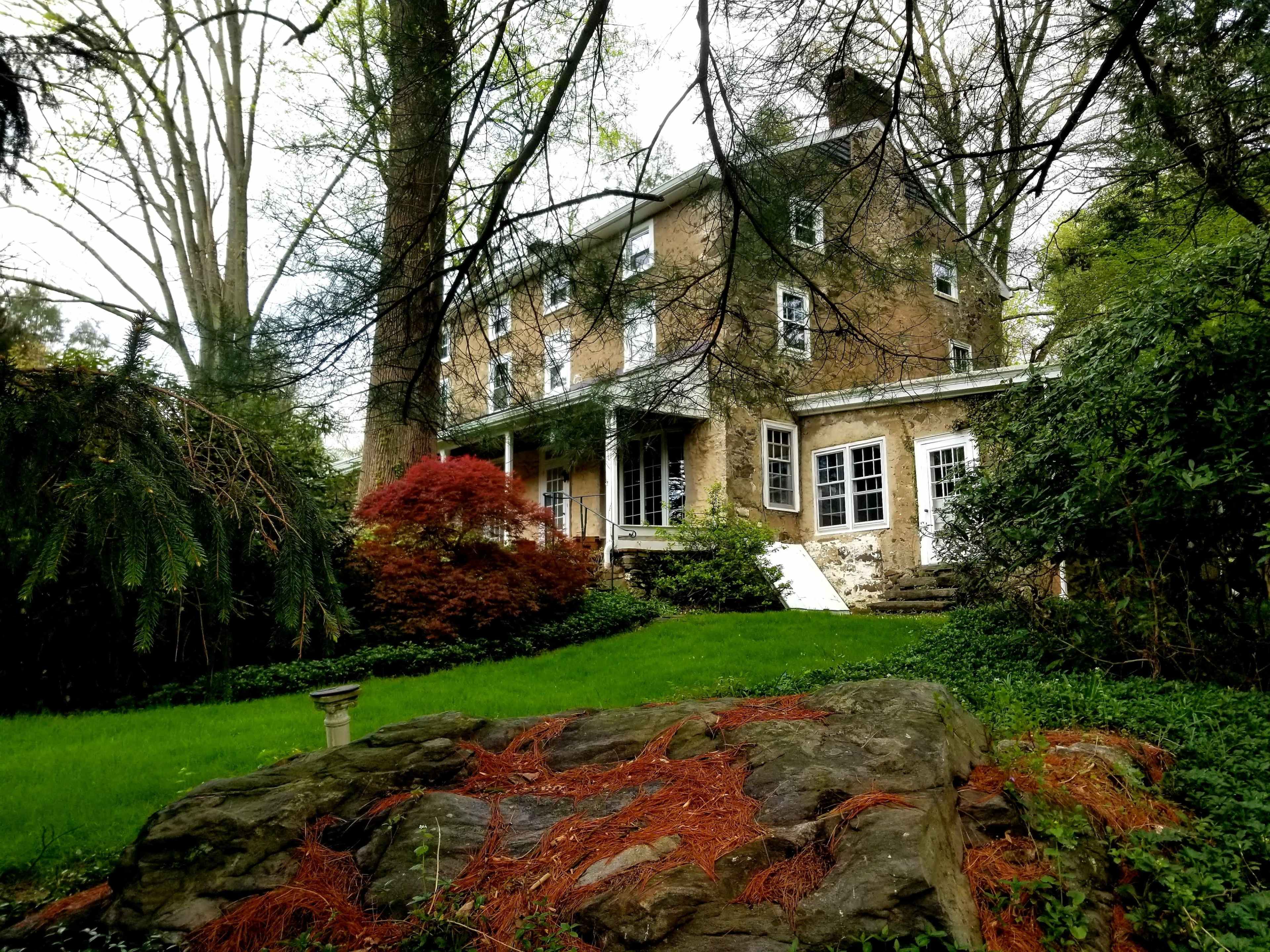 A three-story stone house is surrounded by trees and green foliage, with a red-leafed shrub in the foreground and a stone outcropping nearby.